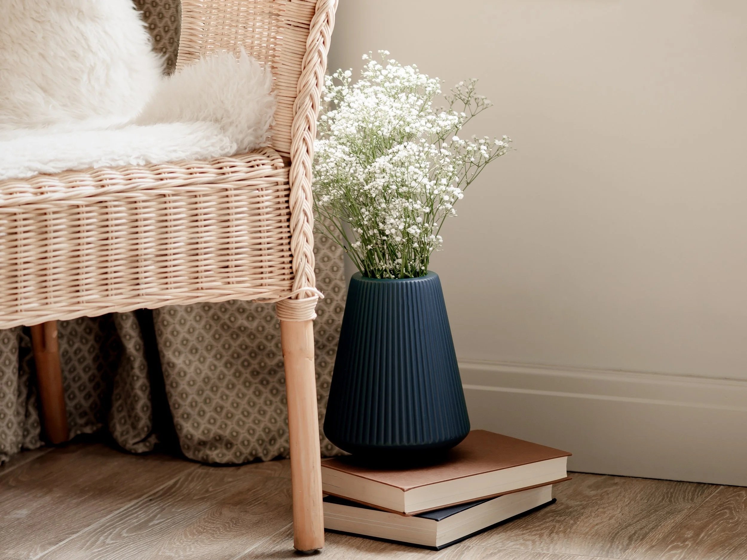 A wicker chair with a furry cushion, a patterned fabric behind it, a blue vase with white baby's breath flowers, and two stacked books on the floor.