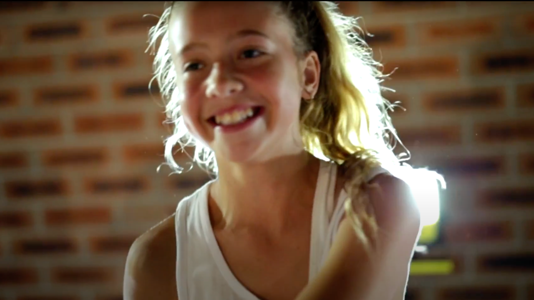 A young woman with blonde curly hair smiling and leaning forward, with a brick wall in the background.