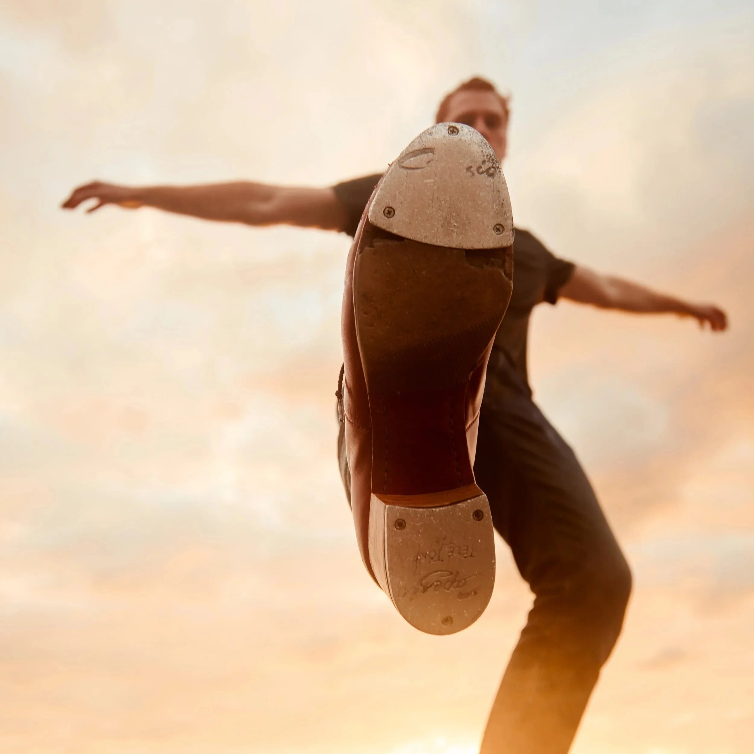 Close-up of a person's foot wearing a brown shoe, with arms extended outward in a wide stance, against a cloudy sky background.