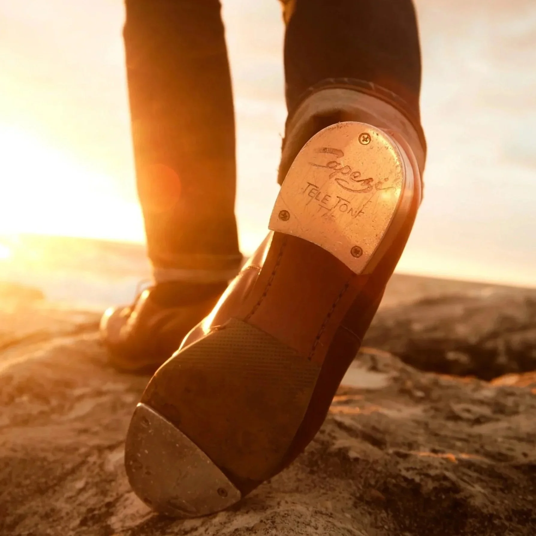 A close-up of a leather cowboy boot with a metal plate on the heel that reads 'Laredo Tele Tone.' The boot is on sandy ground with a sunset or sunrise in the background.