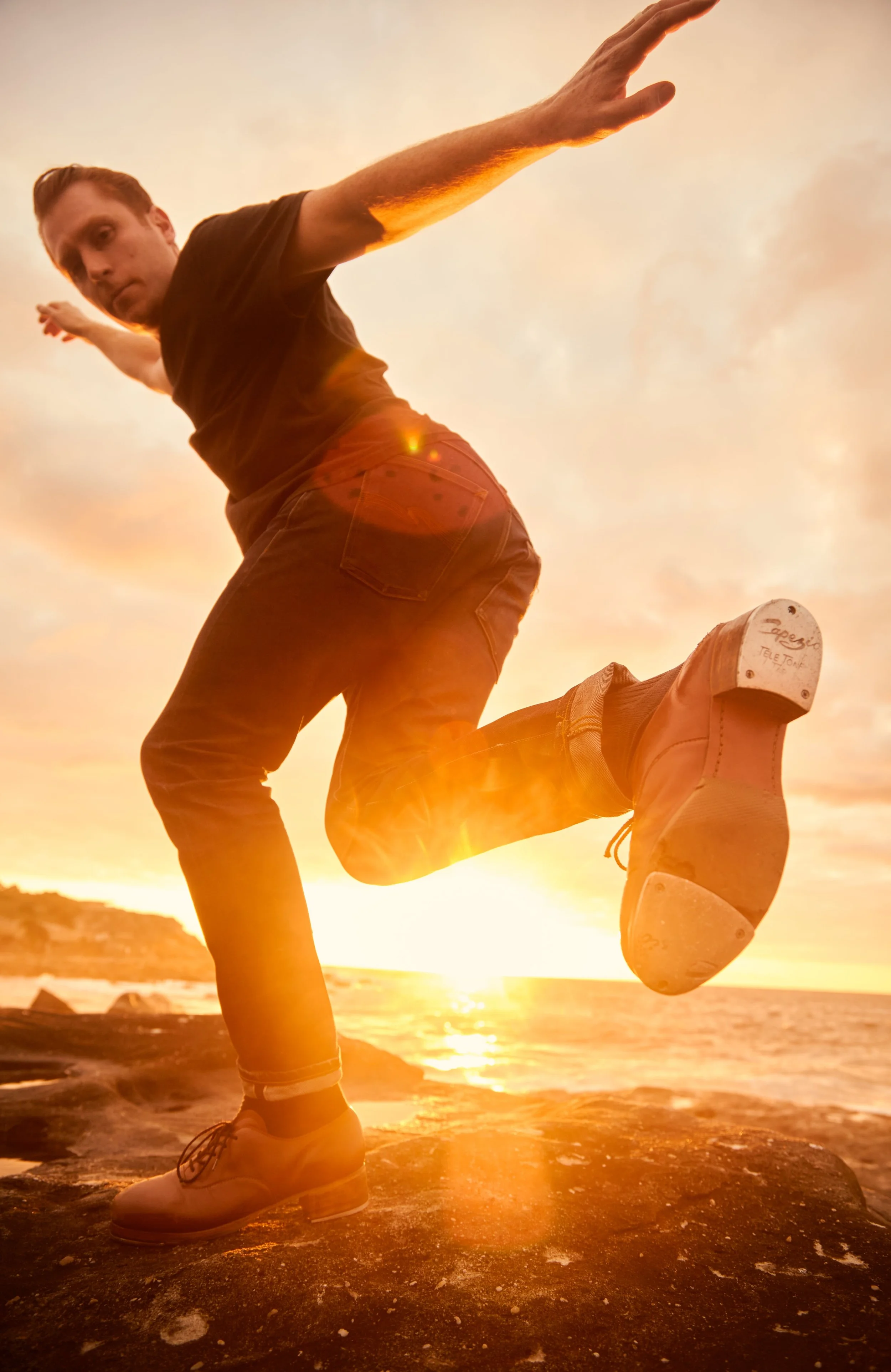 Person performing a one-armed handstand on rocks by the beach during sunset, with one leg bent and the other extended, wearing white sneakers and jeans.