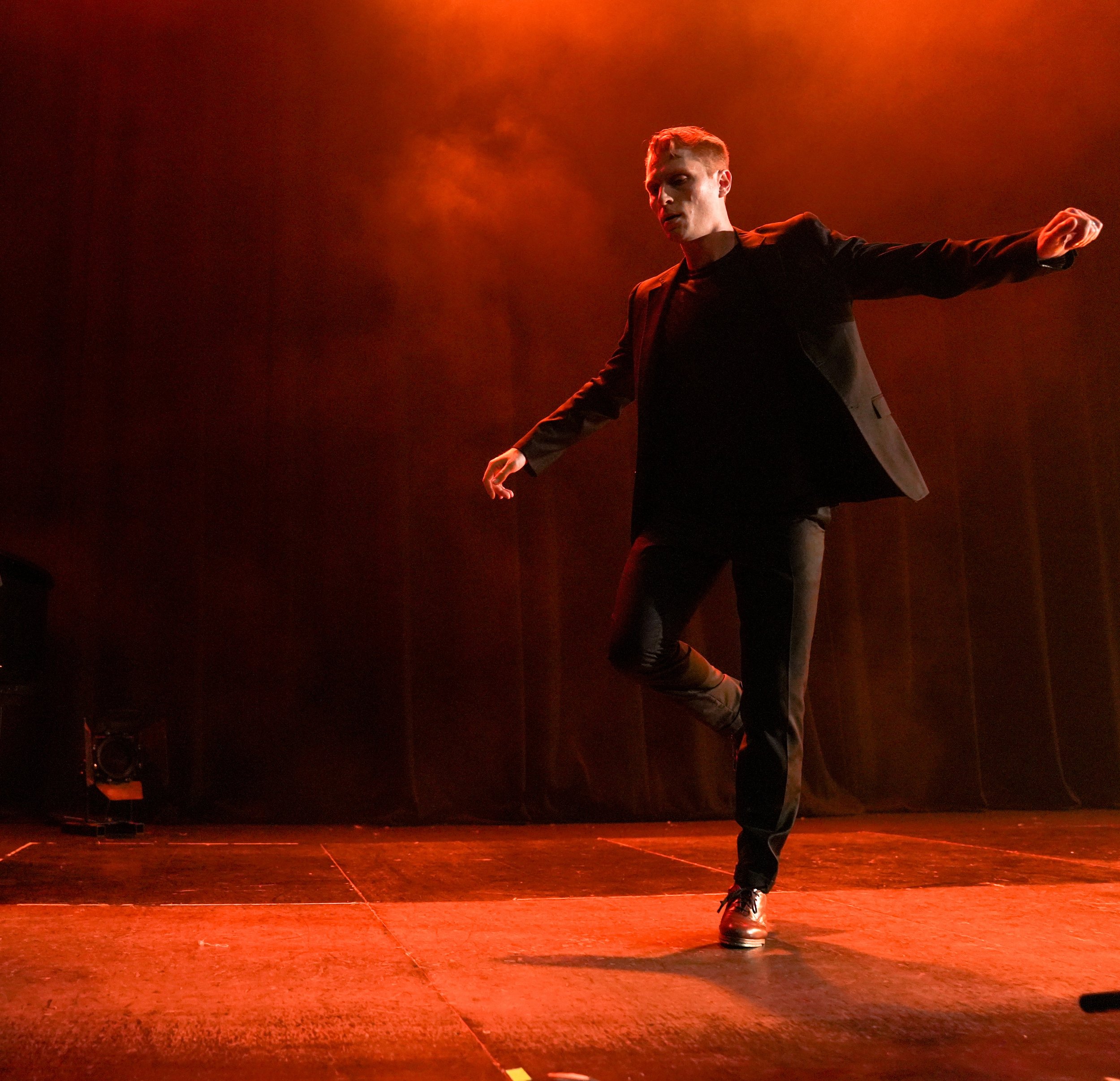A person performing a dance move on a stage illuminated by red lighting, wearing a black blazer, dark pants, and black shoes.