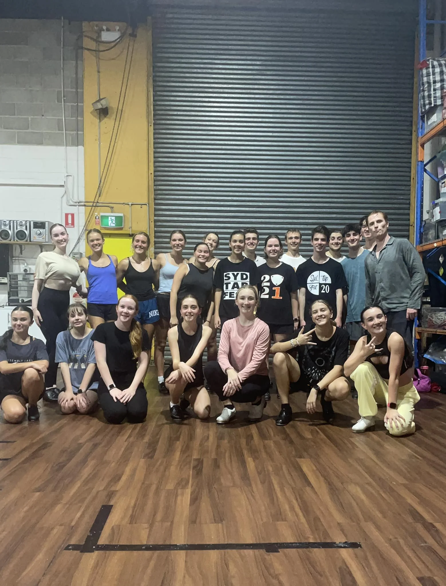 A group of young people, mostly women, posing for a photo inside a gym or dance studio. They are standing and kneeling on a wooden floor, with a large metal roll-up door behind them. Some are smiling and making peace signs, and a man is standing on t