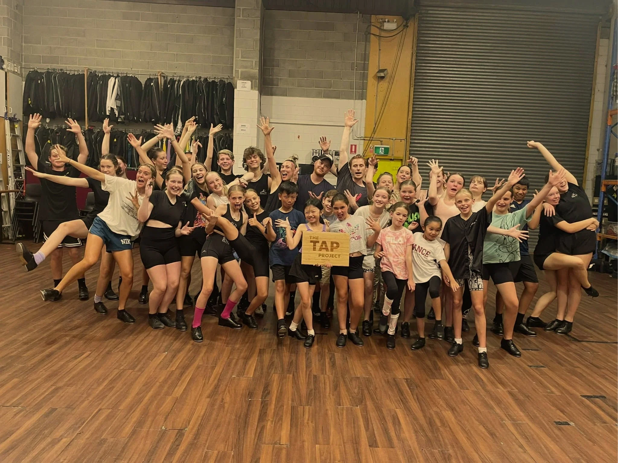 Group of children and teenagers gathered in a gym or dance studio, posing with smiles and various poses, some holding a wooden sign that reads 'The TAP Project.' In the background, there are racks of jackets, bags, and storage shelves.