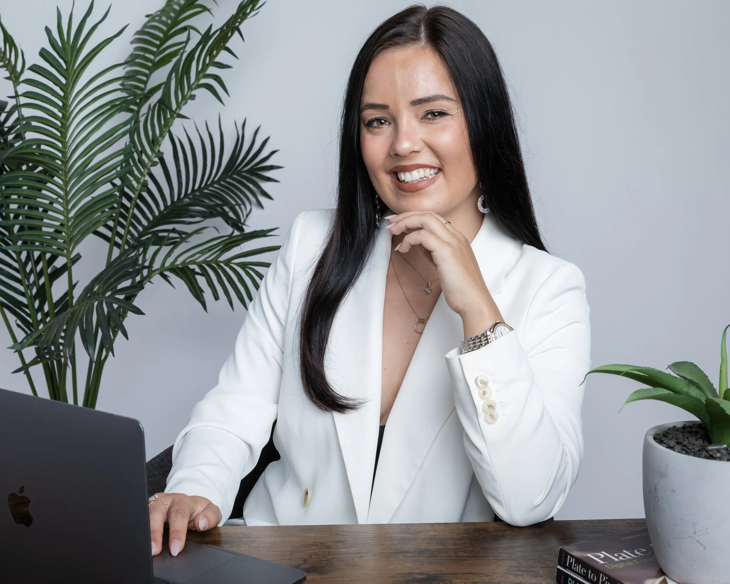 A woman with long dark hair, wearing a white blazer and jewelry, sitting at a desk with a laptop, surrounded by green plants in a bright, professional setting.