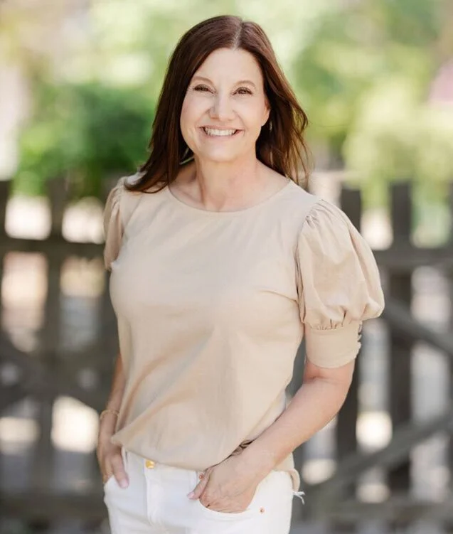 A woman with shoulder-length brown hair, smiling, wearing a beige top with puffed sleeves and white pants, standing outdoors on a sunny day with greenery in the background.