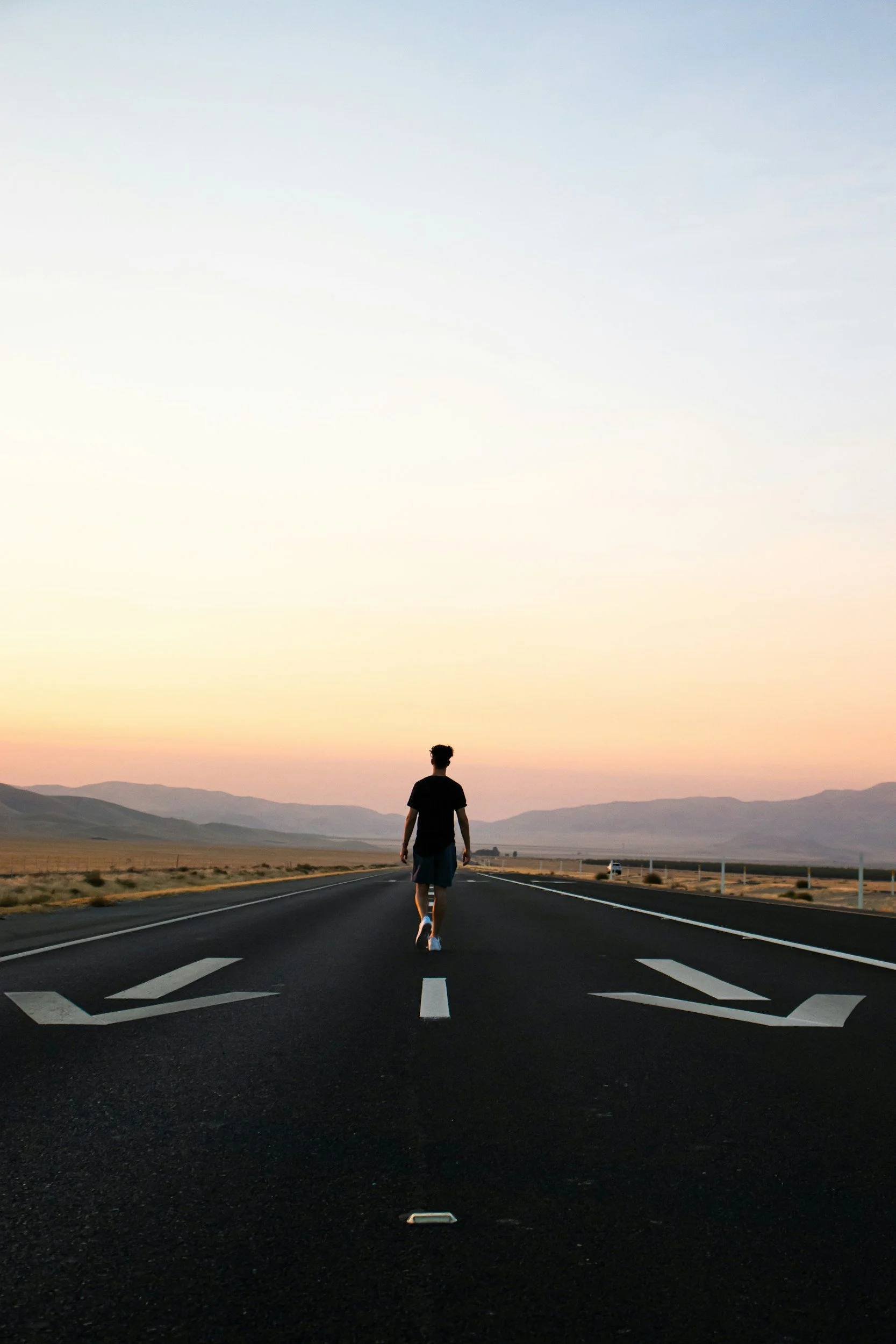A person walking on a deserted road in a vast open landscape with mountains in the distance during sunset.