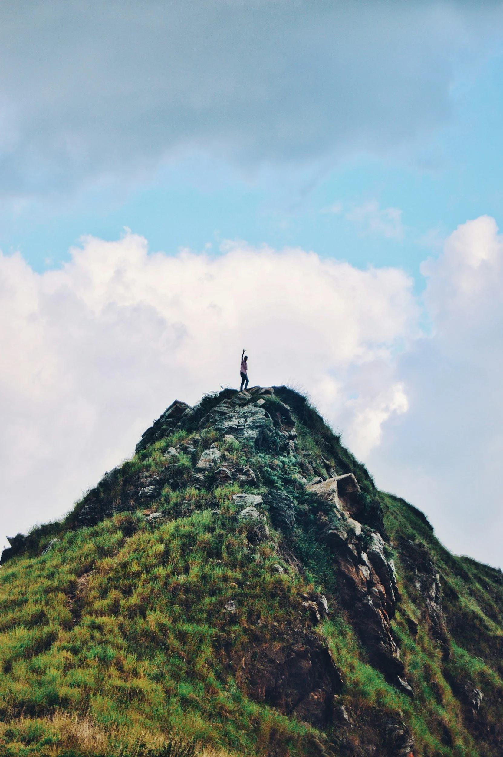 Person standing on a rocky mountain peak under a cloudy sky, raising one arm in the air.