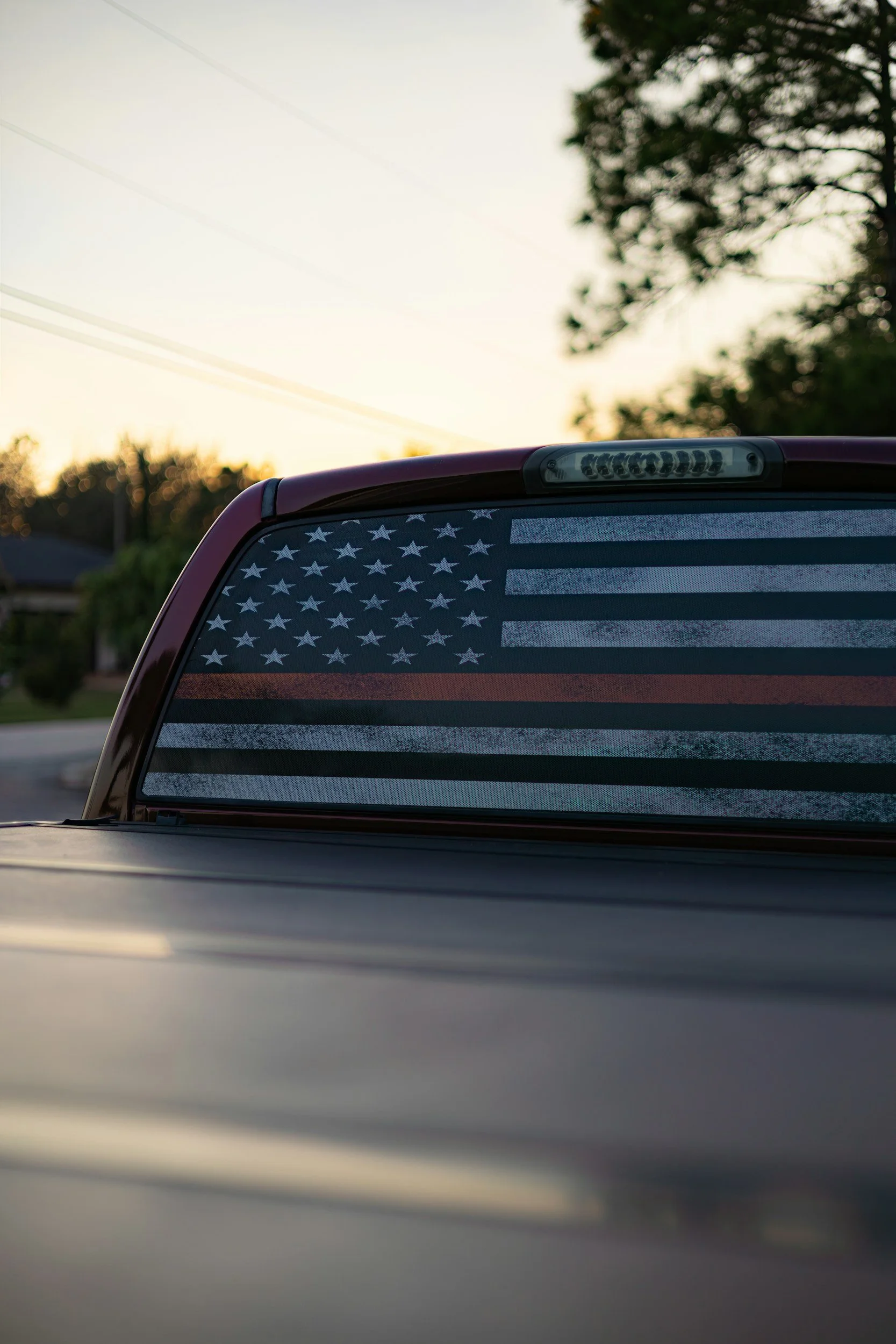 The back window of a vehicle decorated with an American flag sticker, parked outdoors during sunset, with trees and power lines in the background.