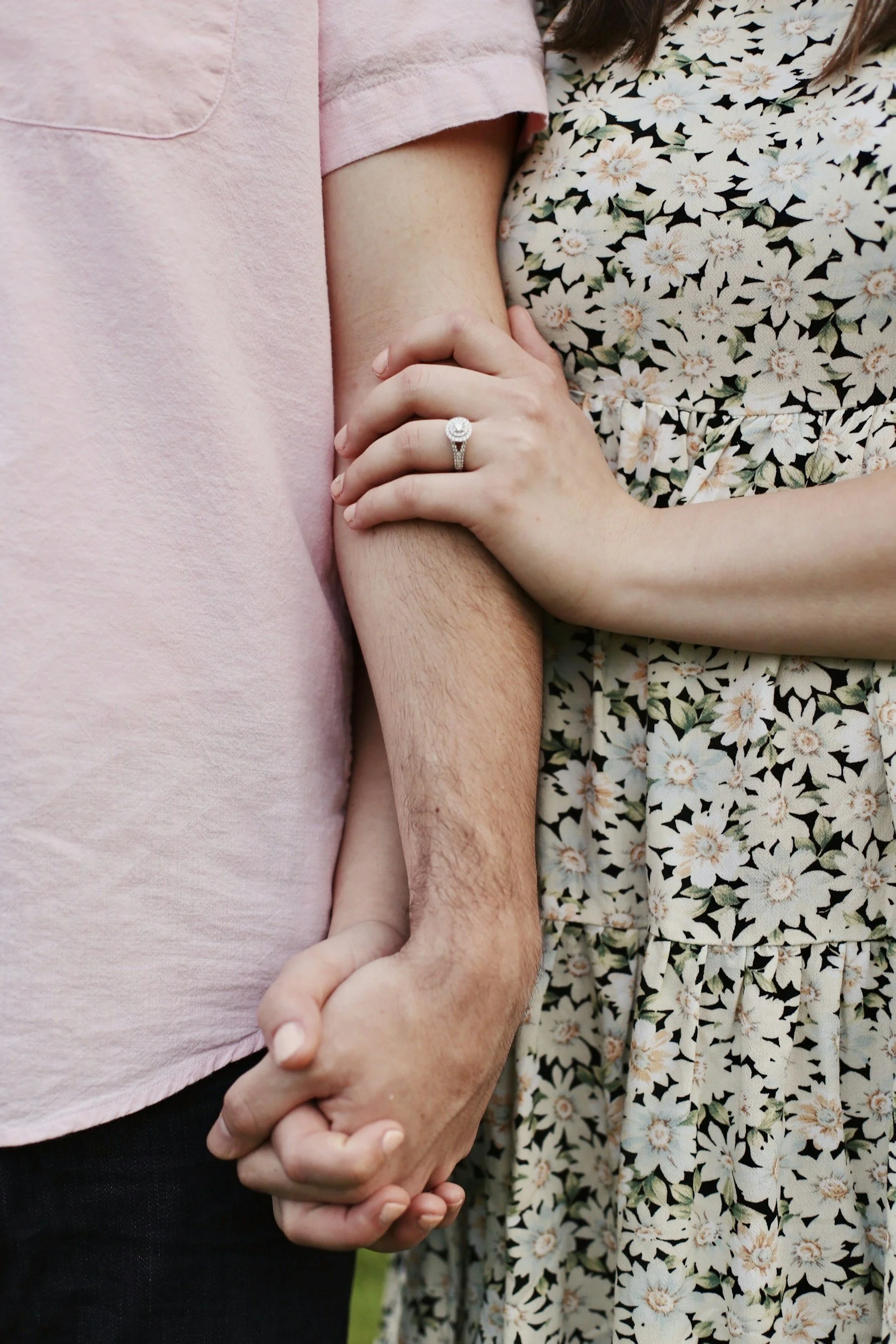 A close-up of two people holding hands, one person wearing a pink shirt and another person wearing a floral dress, with one wearing a diamond ring.
