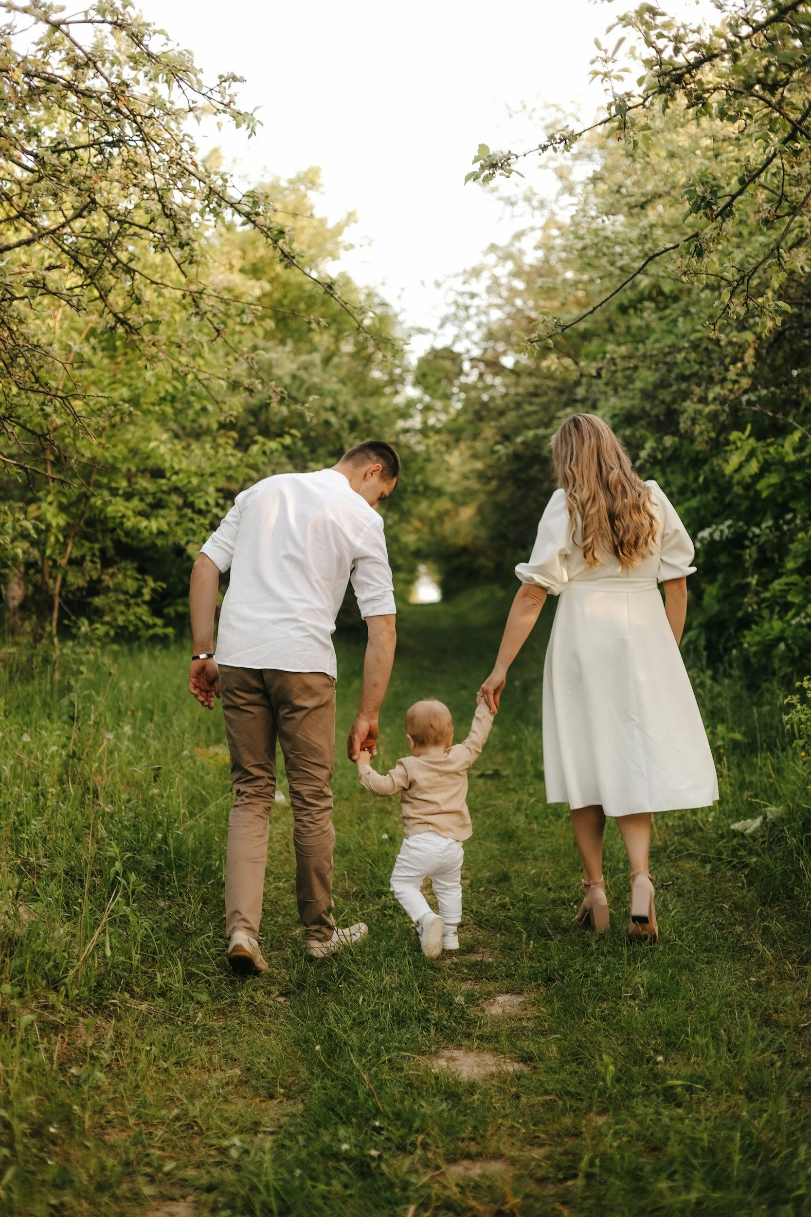 A family of four, including a man, a woman, and two small children, walking hand-in-hand along a grassy forest trail surrounded by trees and greenery.