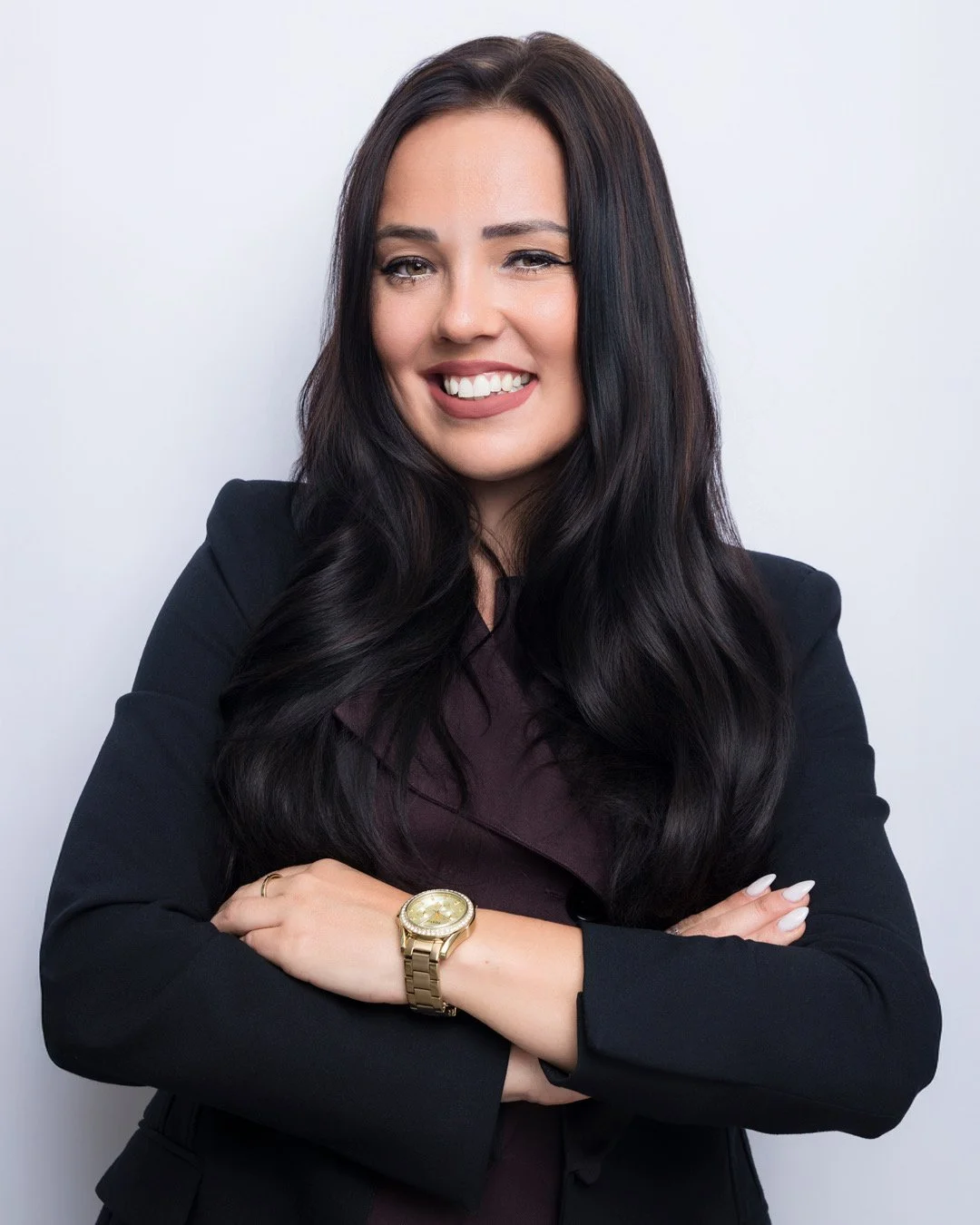 A woman with long, dark hair, wearing a black blazer and maroon shirt, smiling with arms crossed, against a plain white background.