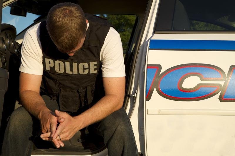 A police officer sitting on the open door of a police vehicle, looking down and clasping his hands together.