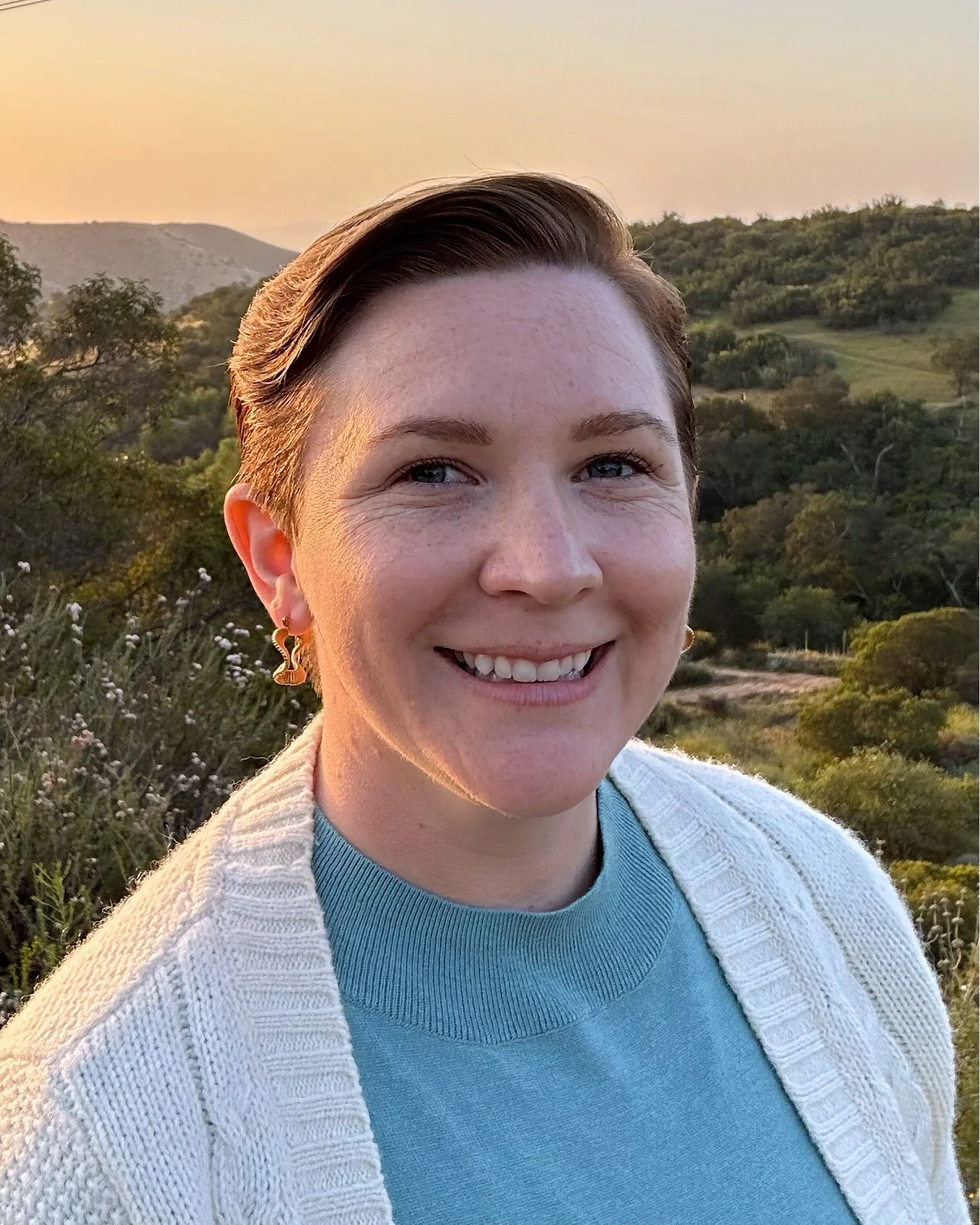 A woman smiling outdoors during sunset with hills and green trees in the background, wearing a blue top and a cream-colored cardigan.