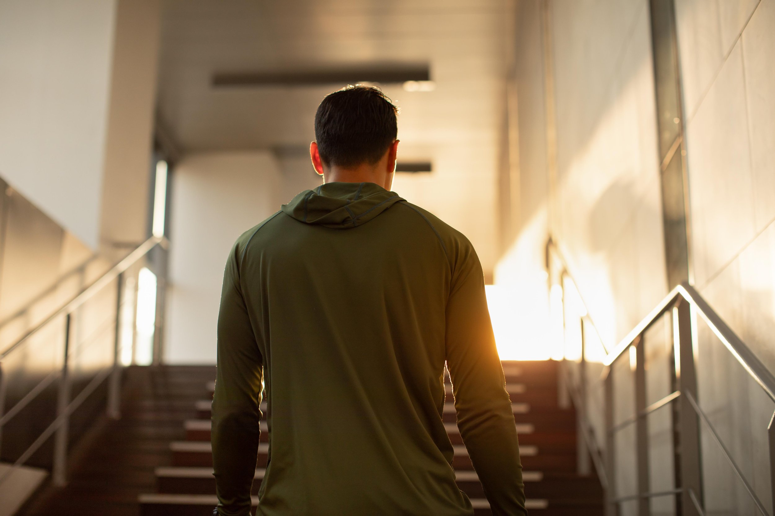 A person with short hair wearing a green jacket walking up stairs in a corridor with sunlight illuminating the space.
