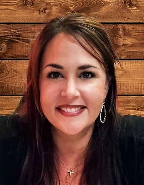 A smiling woman with long dark hair, wearing earrings and a necklace, in front of a wooden wall.