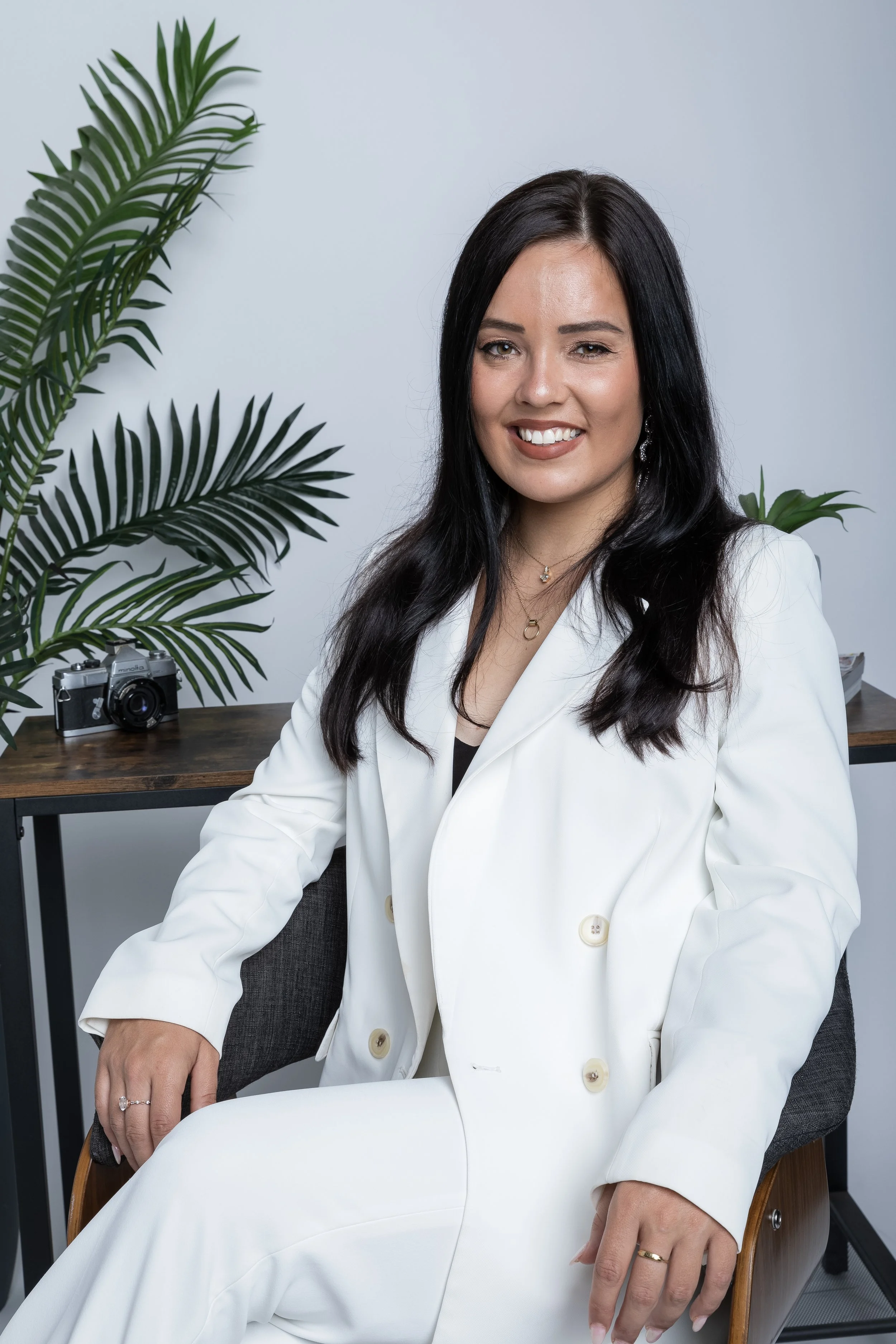 A woman with long black hair, wearing a white blazer and pants, sitting on a wooden chair in an office setting with green plants and a vintage camera on a desk behind her.