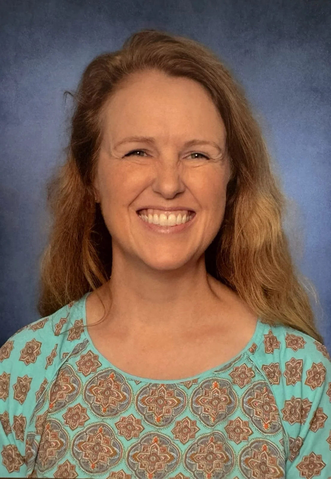 A woman with long wavy red hair smiling, wearing a turquoise patterned top, standing against a blue background.