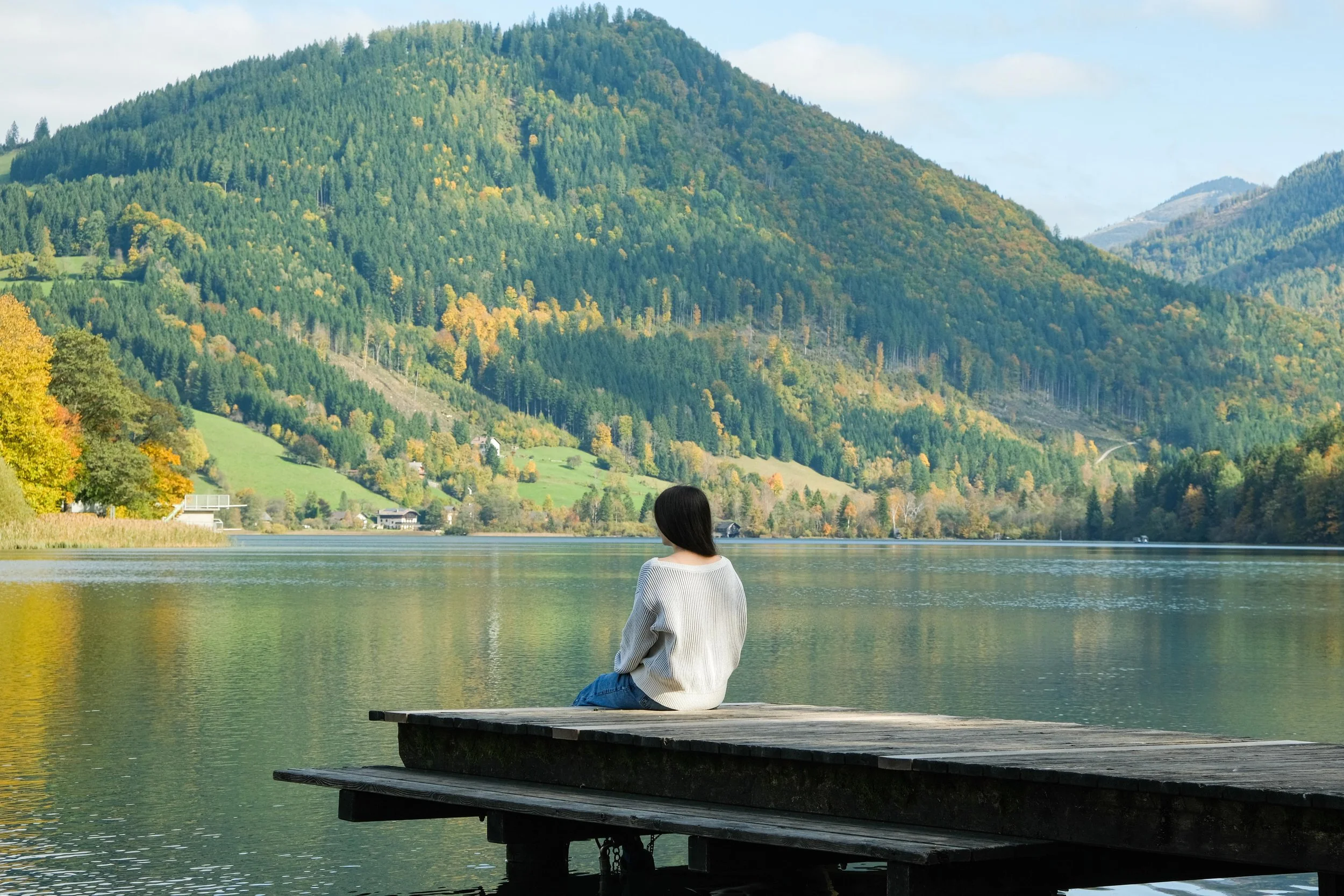 A person sitting on a wooden dock by a lake, with view of lush green mountains and a partly cloudy sky in the background.