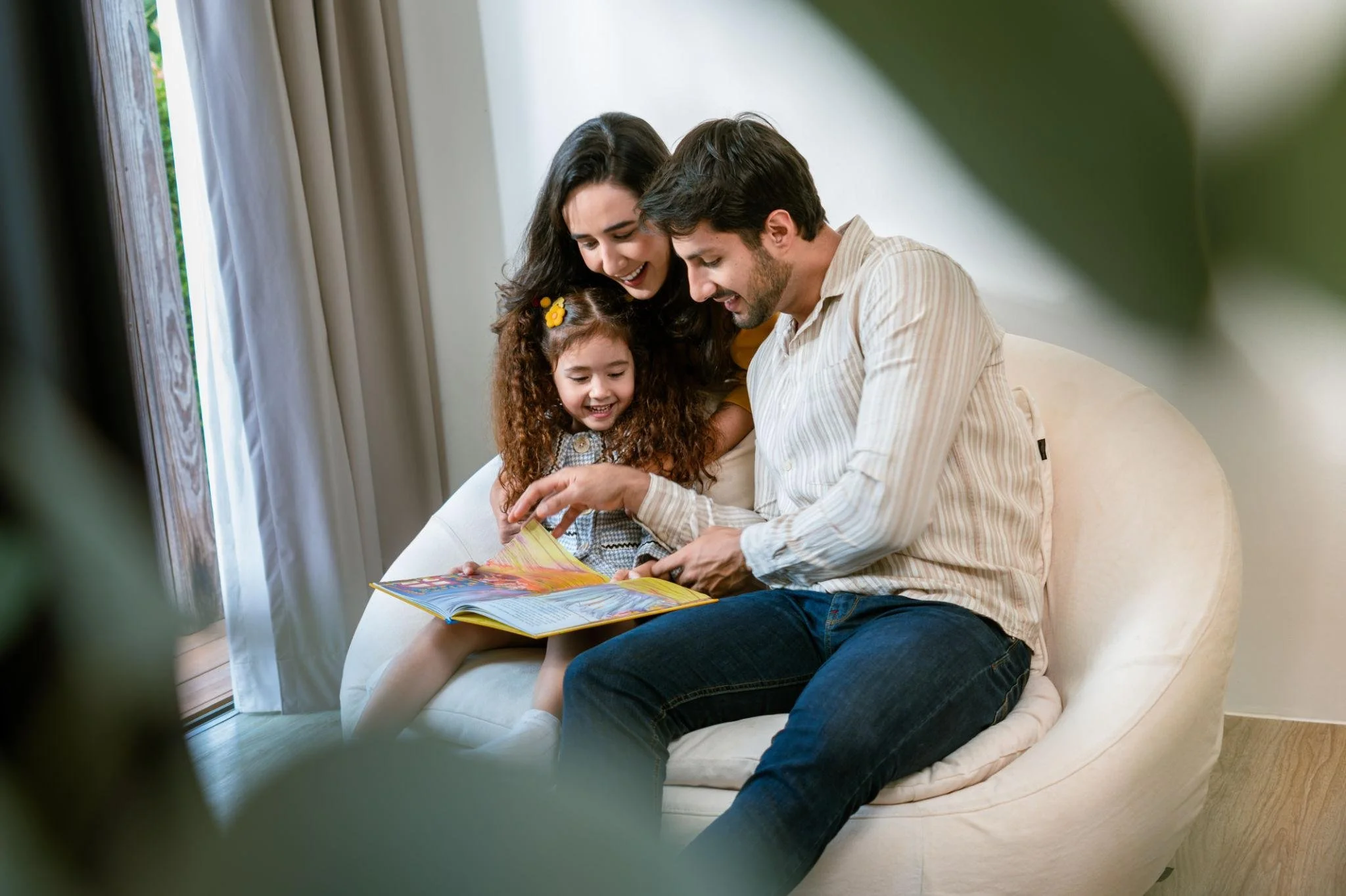 A family of three, a man, a woman, and a young girl, sitting on a white couch and looking at a colorful book together, smiling happily in a bright room near a large window with white curtains.