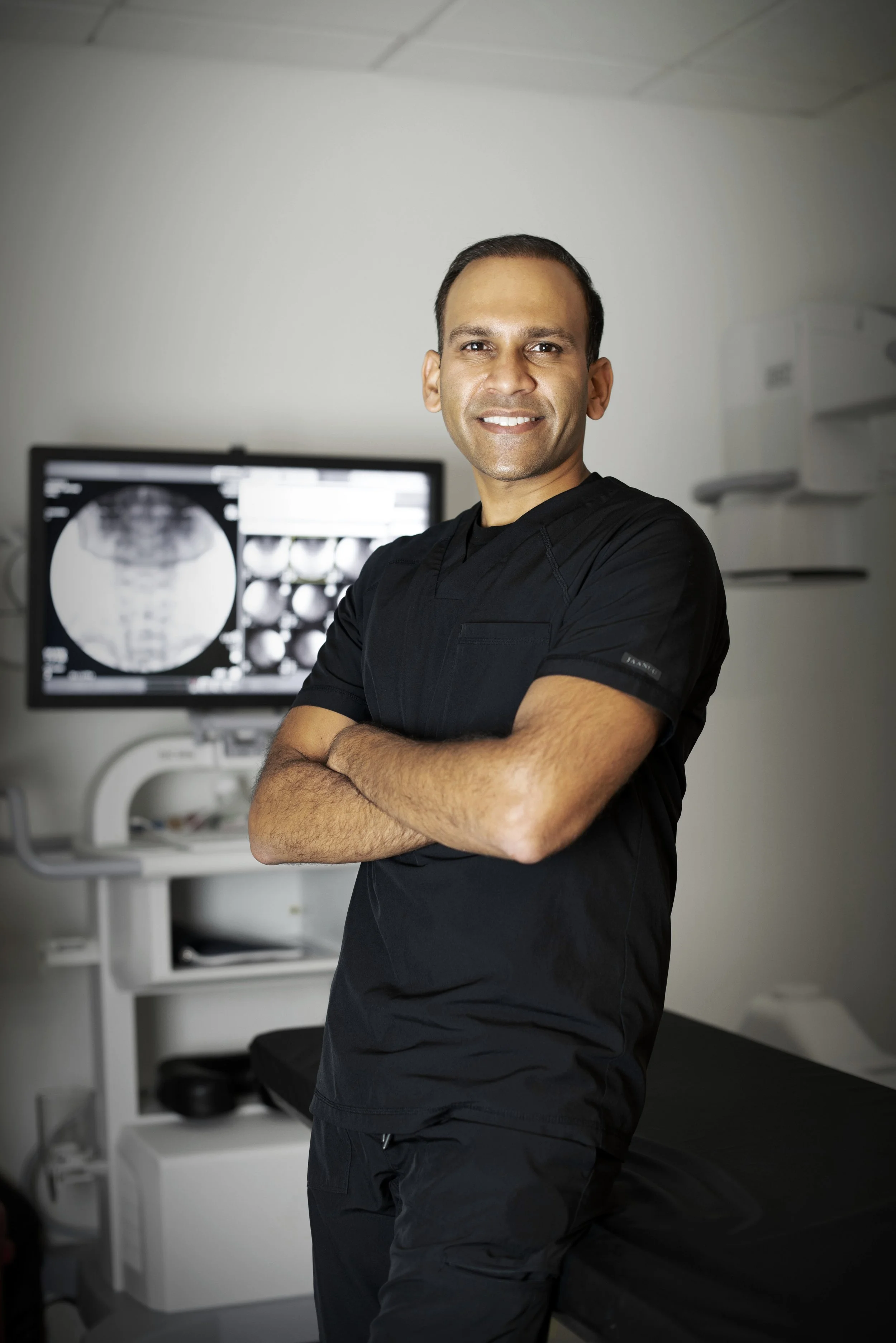 A smiling male healthcare professional standing with arms crossed in a medical office, with medical imaging displayed on a monitor behind him.