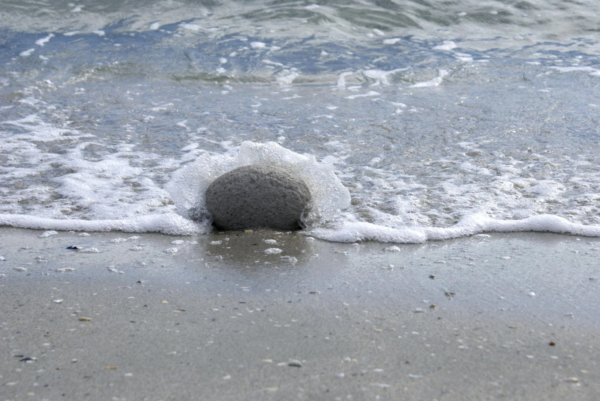 A large rock on the sandy beach near the shore, with small waves washing around it.