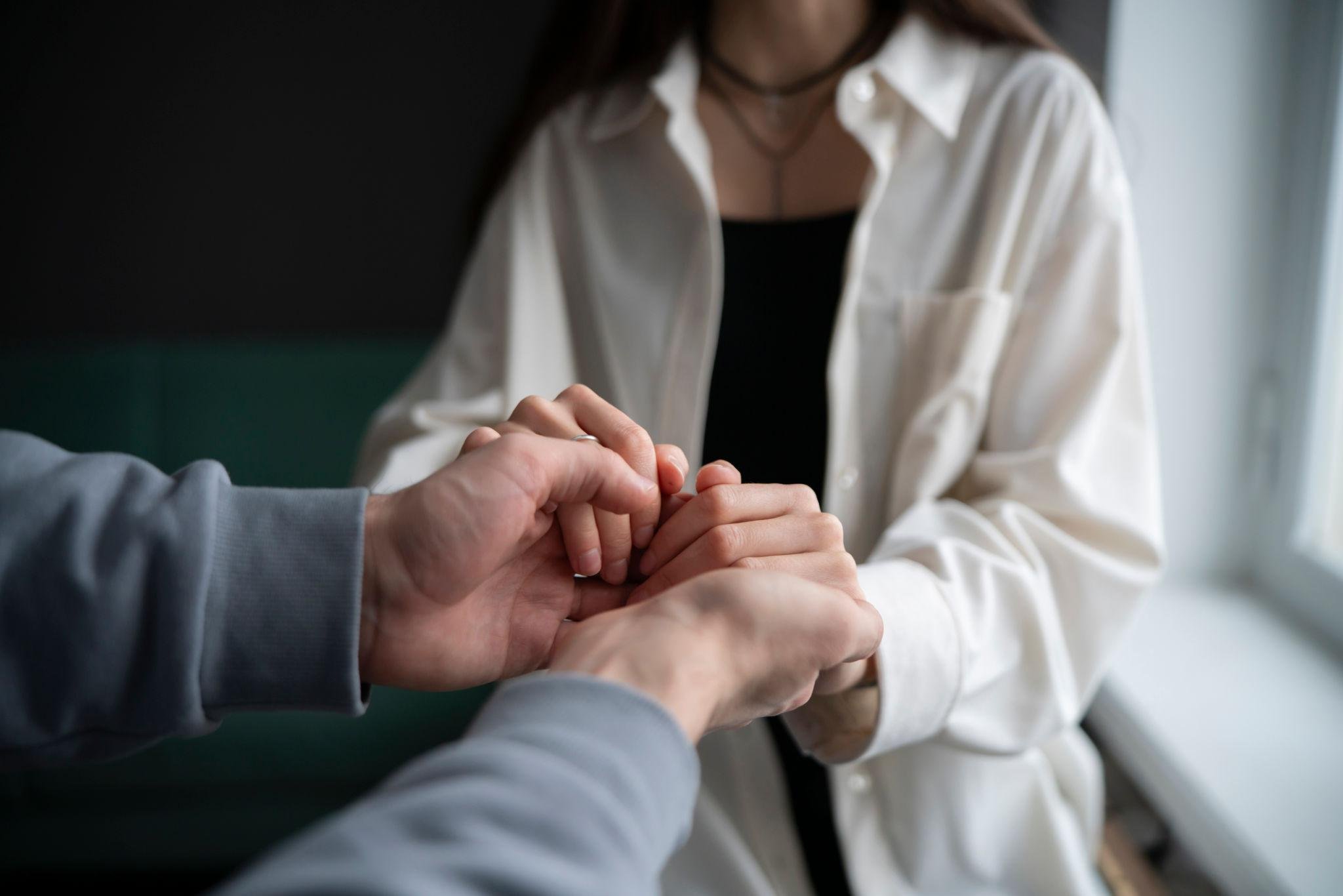 Close-up of two hands holding each other, with a woman in a white shirt and a black choker necklace slightly blurred in the background.