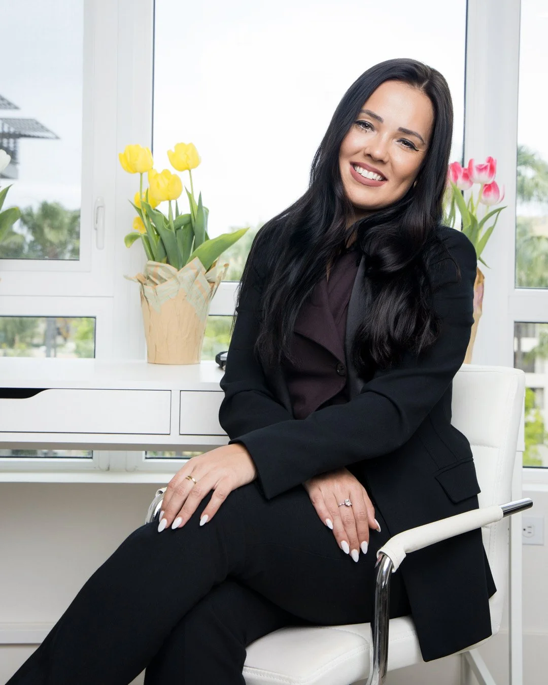 A woman with long dark hair, sitting on a white chair in front of a white desk with yellow and pink tulips in pots, smiling at the camera.
