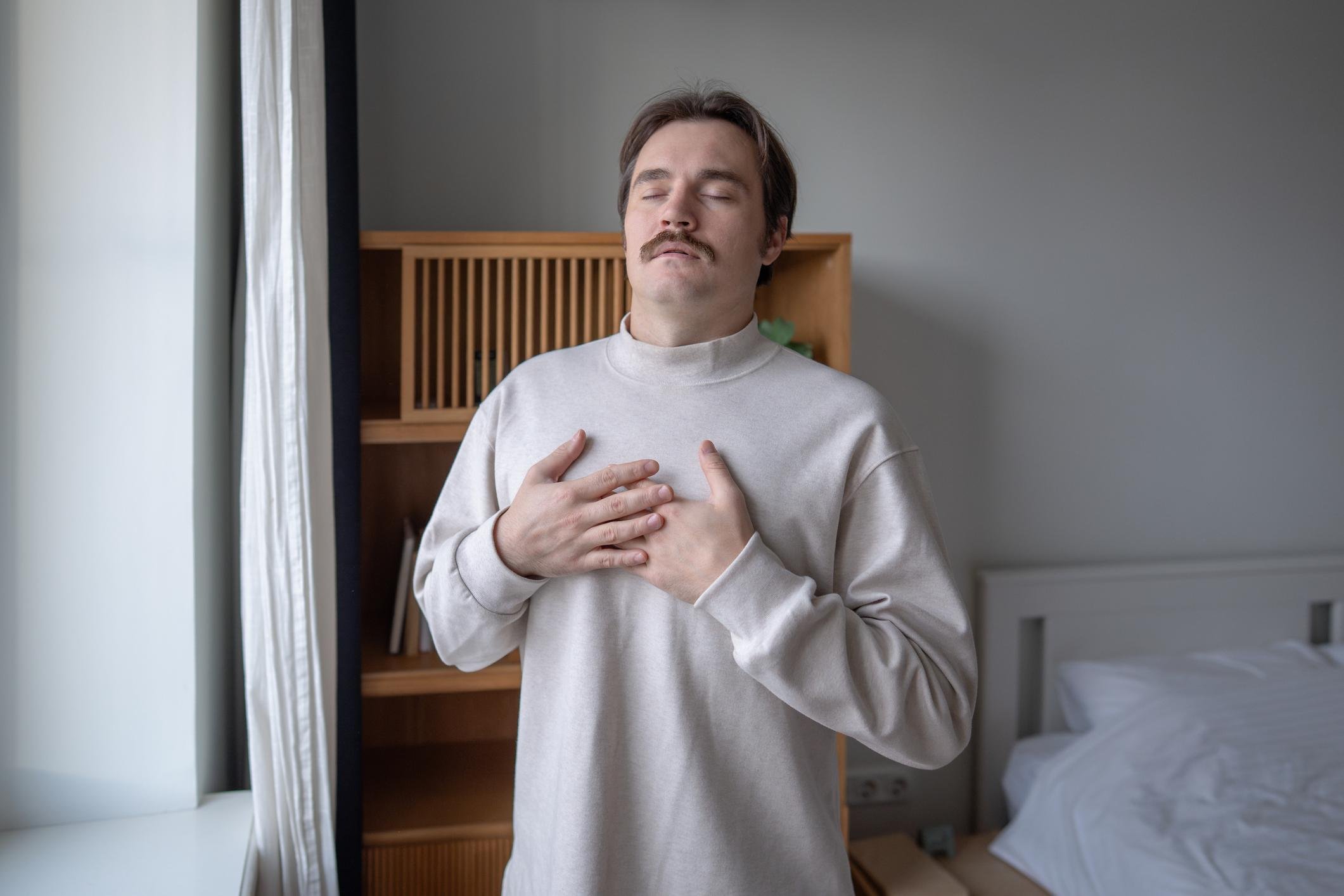 A man with a mustache and brown hair, wearing a beige sweater, is standing indoors with his eyes closed and his hands on his chest, appearing to meditate or relax.