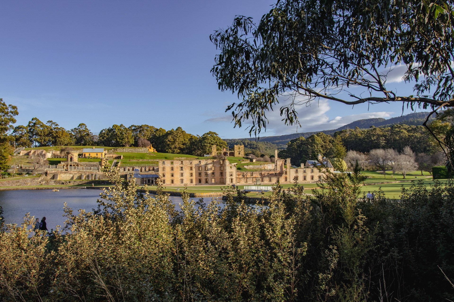 A scenic landscape featuring the historic Port Arthur building complex, a body of water in the foreground, lush green trees, and hills in the distance under a clear blue sky.