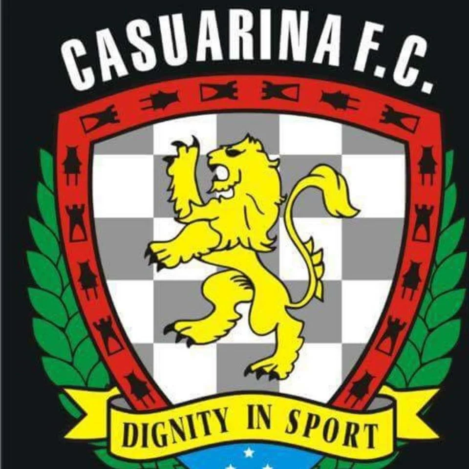 The emblem of Casuarina F.C. features a yellow lion on a checkered gray and white background, encircled by a red border with black tower symbols, and green laurel leaves on either side. The team motto, 'Dignity in Sport,' is on a yellow banner at the bottom.