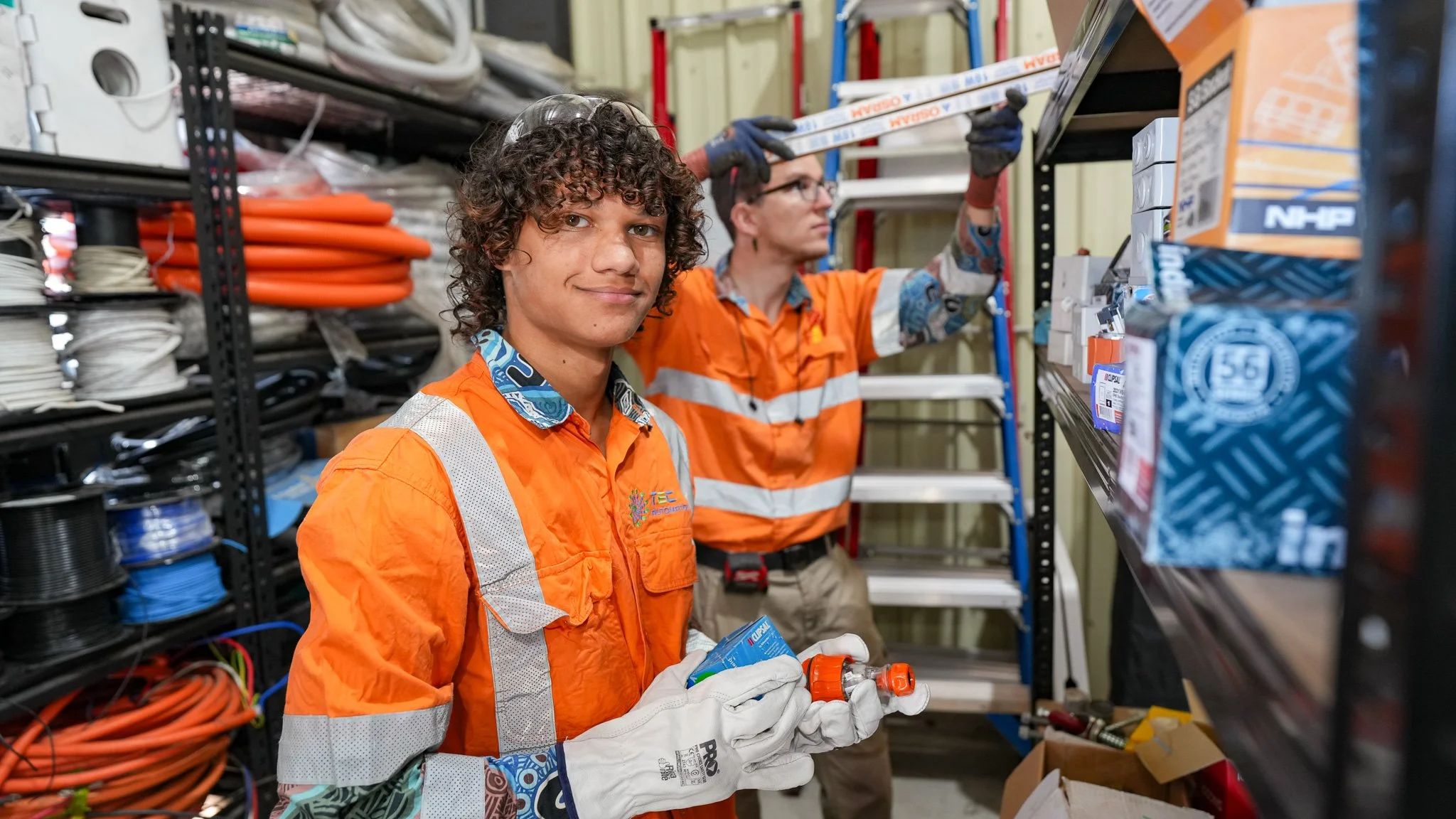 Two TEC Automation workers in orange safety uniforms shop for electrical supplies in a hardware store, with shelves of wires and tools around them.