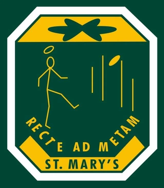 A shield-shaped emblem with a dark green background, featuring a yellow play symbol, a dancing stick figure with a halo, and falling balls. The words "RECEDE AD METAM" are written in yellow, and at the bottom, it says "ST. MARY'S" in bold yellow letters.