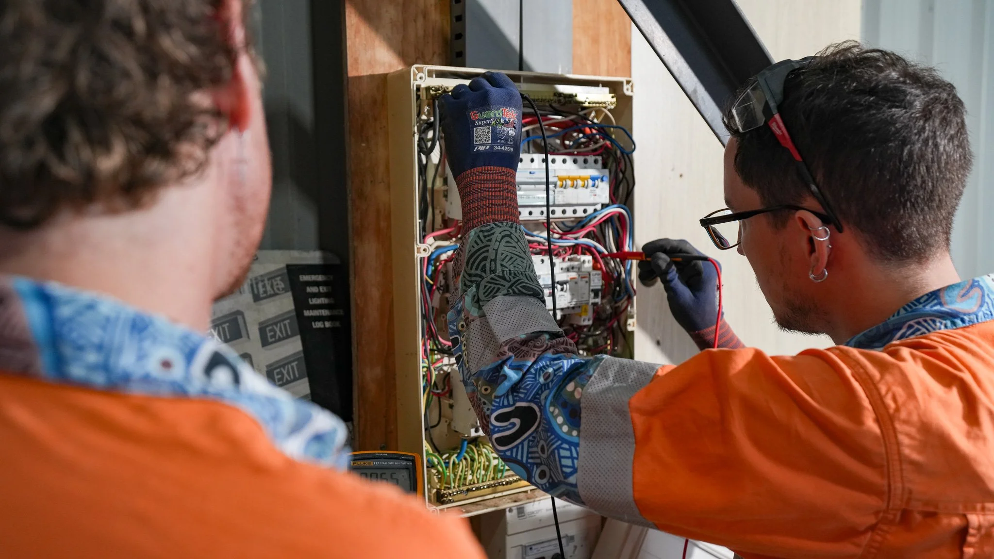 Two TEC Automation electricians working on an electrical panel, one with a multimeter, wearing safety gloves, safety glasses, and orange work jackets.