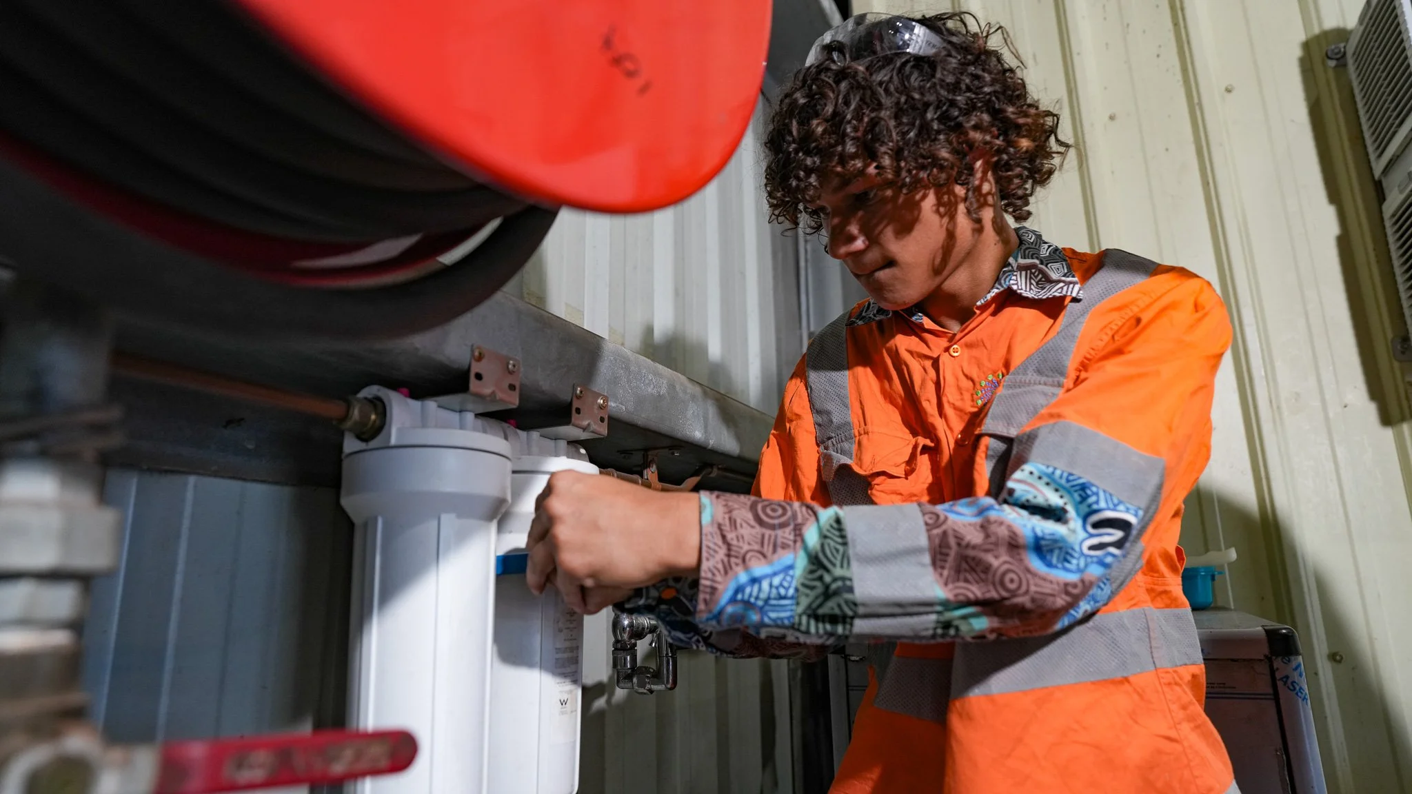 A technician with curly hair wearing sunglasses and an orange safety jacket working on equipment prefixed by plumbing and mechanical components indoors.