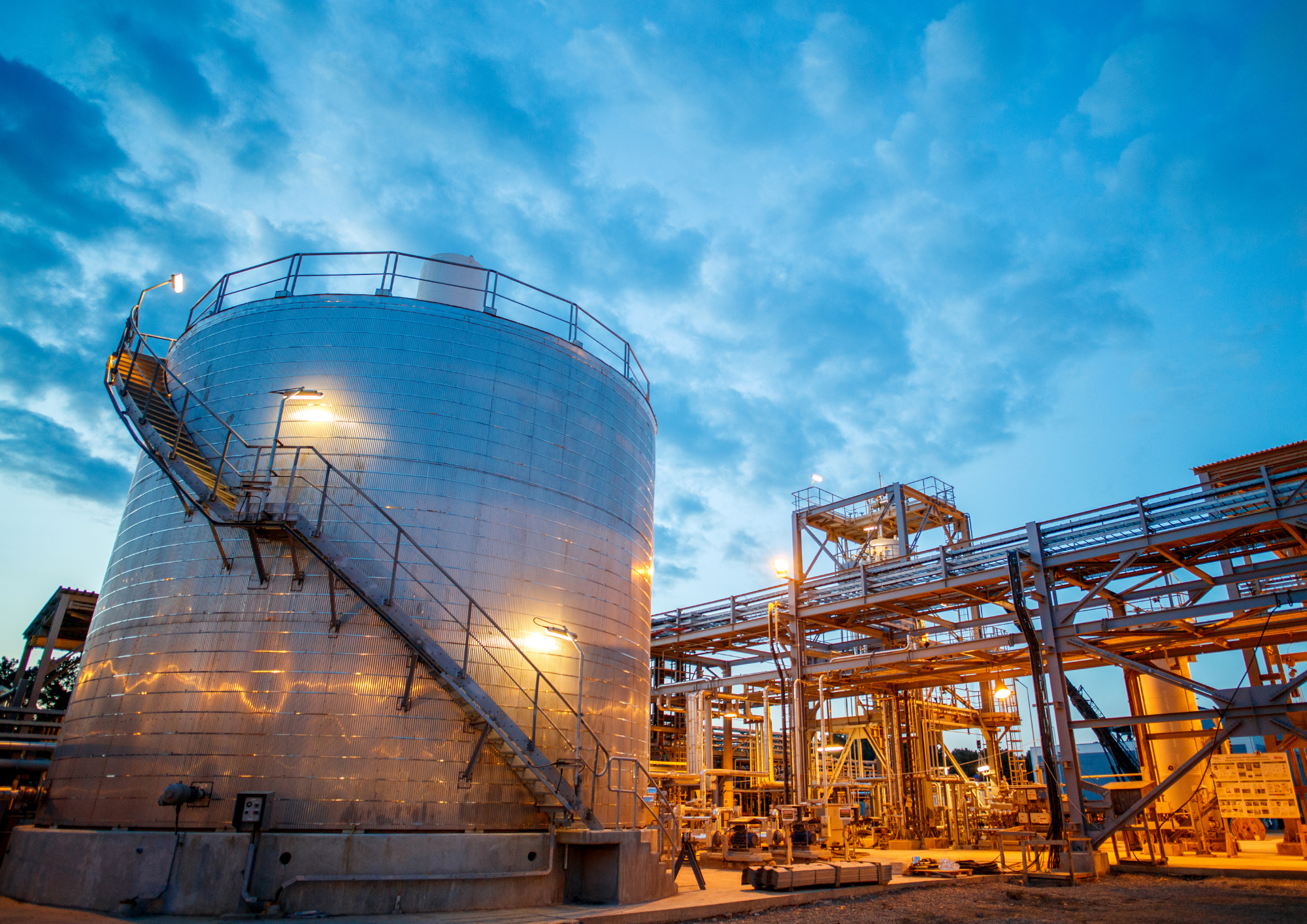 Industrial facility with large metallic tanks and interconnected metal walkways illuminated by warm lighting, set against a twilight sky with scattered clouds.