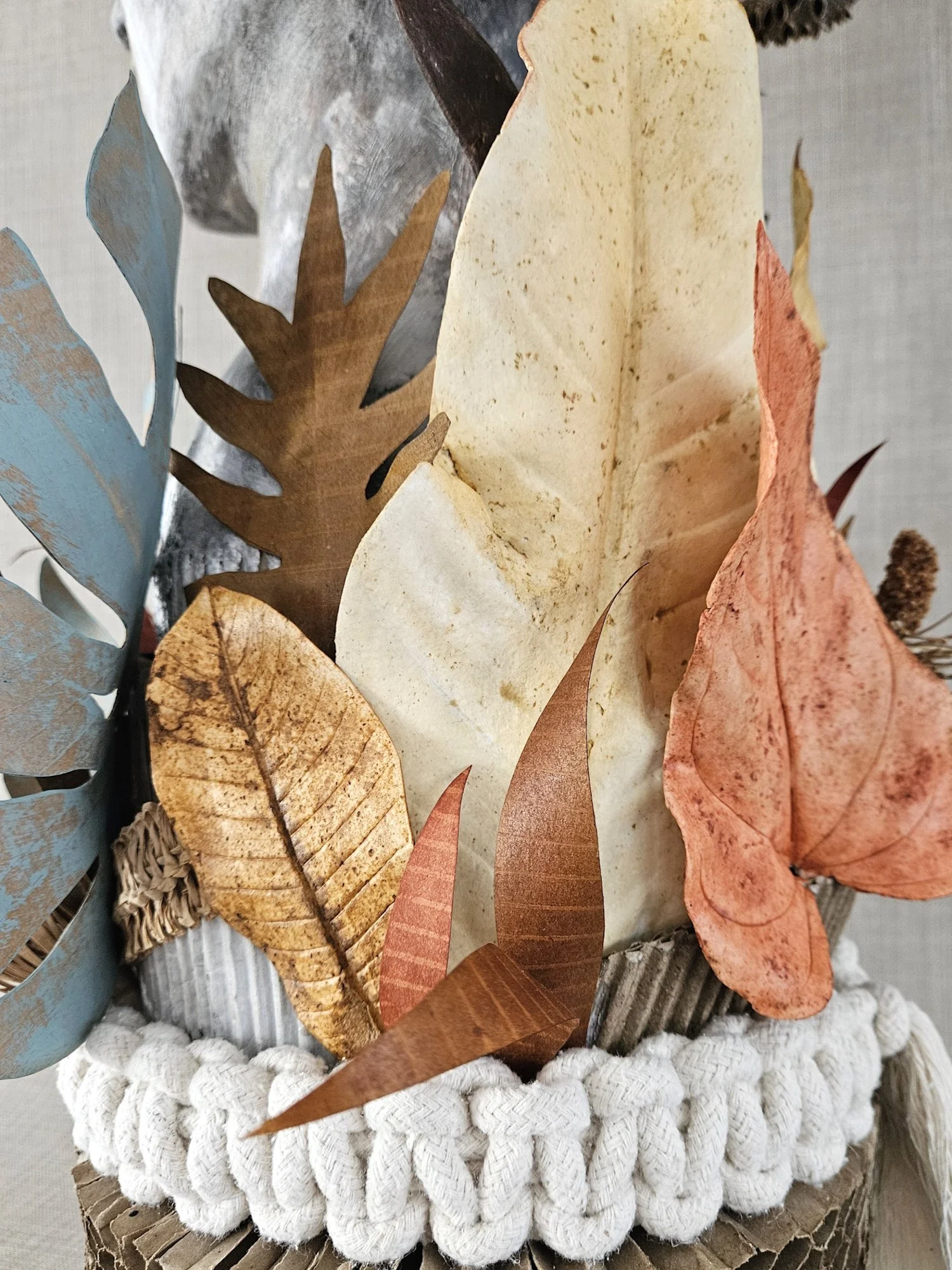 Decorative arrangement of paper leaves in various colors placed on a white, chunky-knit circular base, with a textured gray background.