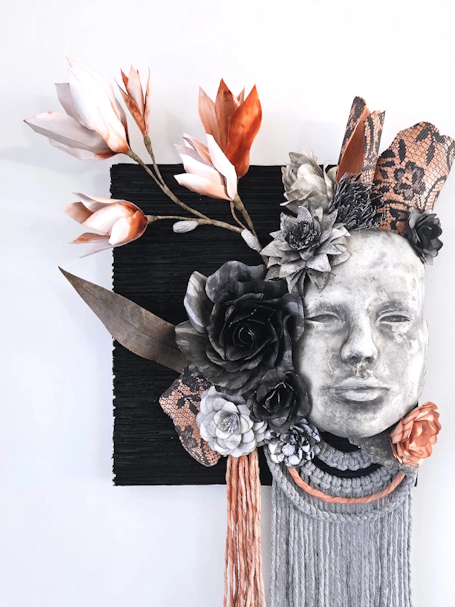 A mixed media artwork combining a paper mache sculpture of a human face with artificial flowers, lace, fabric, and other decorative elements arranged around the head on a black rectangular background.