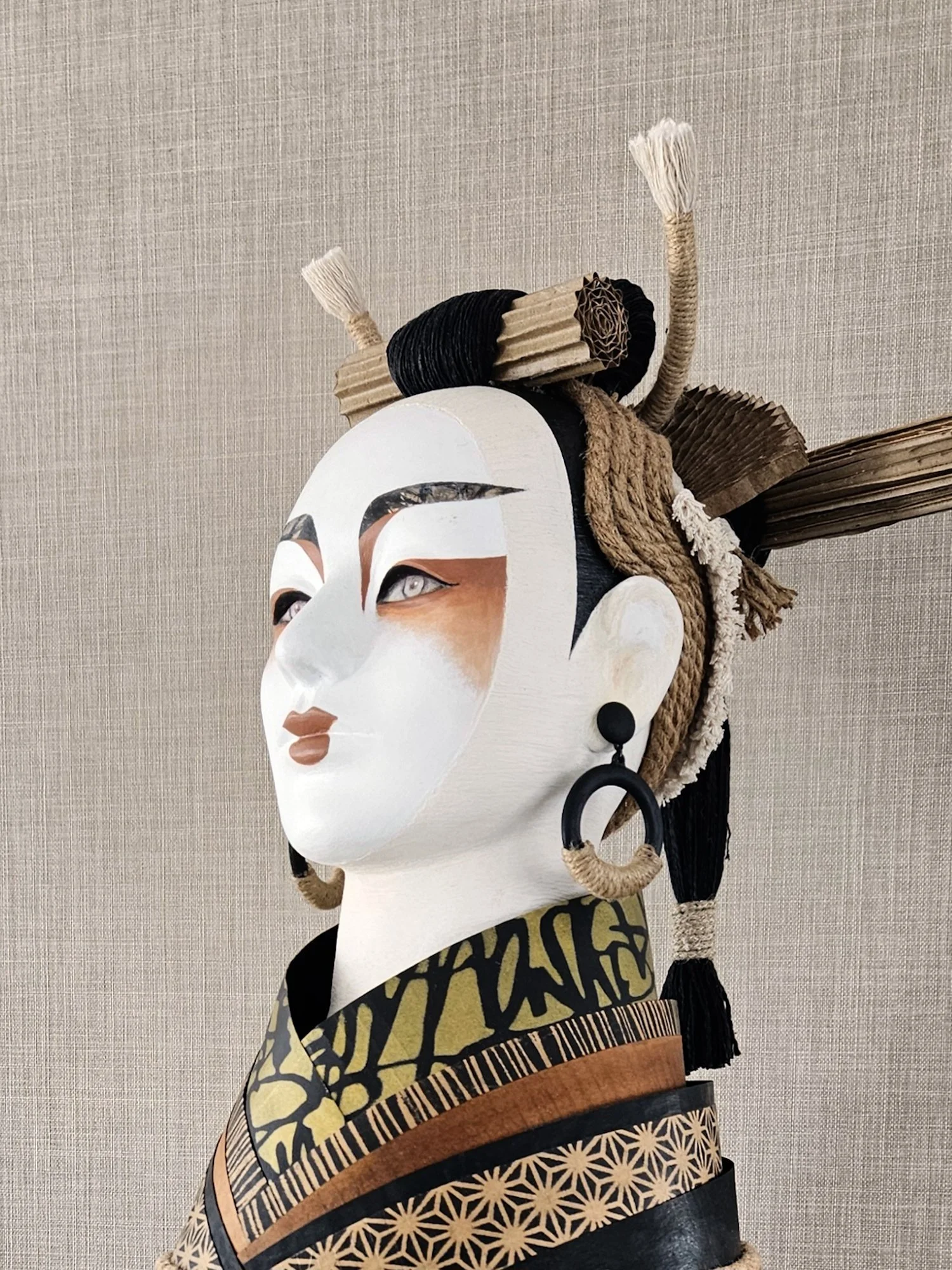 Close-up of an artistic mask with elaborate headdress and intricate designs, featuring a white face with stylized eyes and lips, set against a plain background.