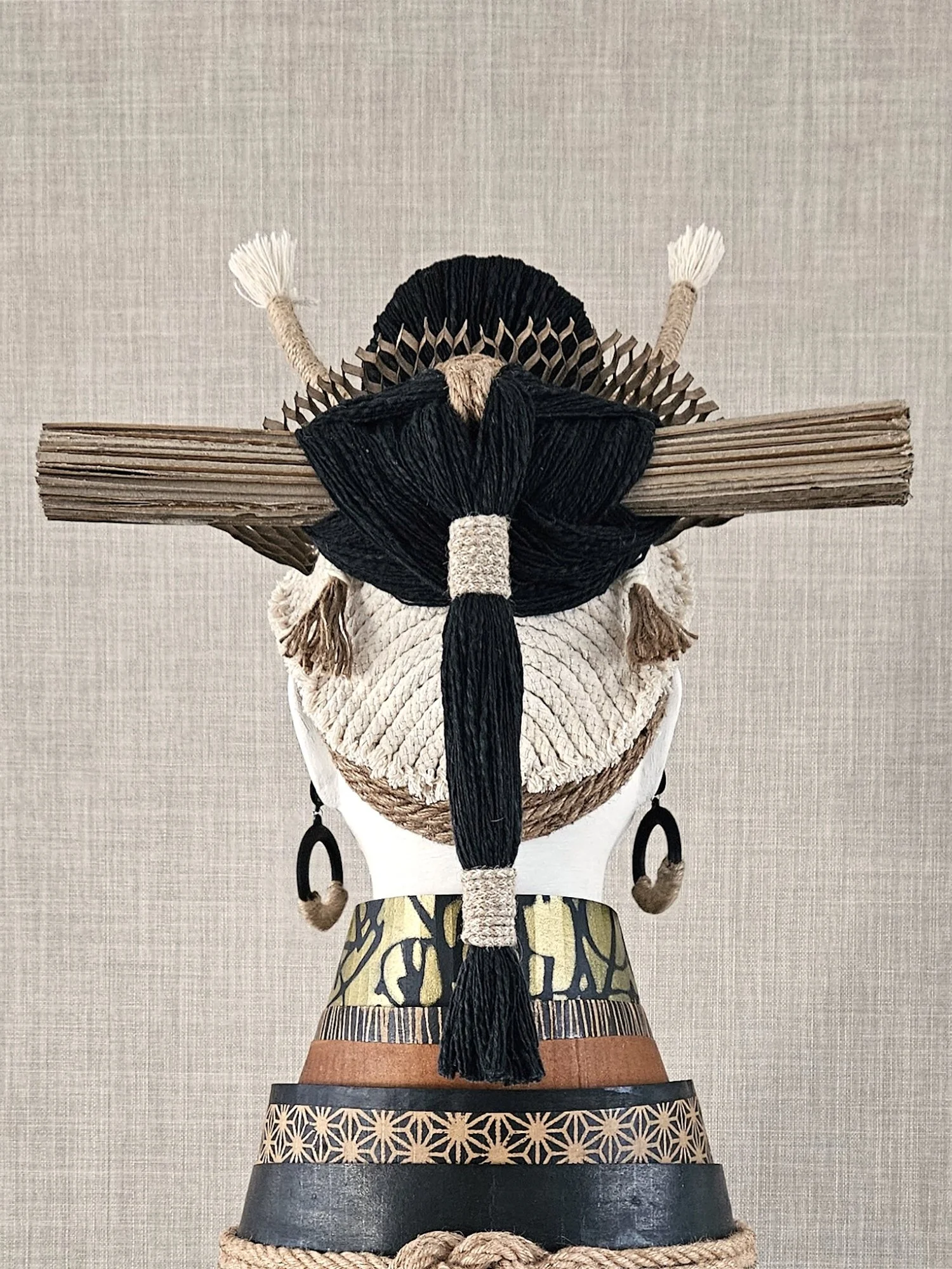 A traditional Japanese doll with intricate clothing, including a black and gold patterned kimono, braided rope details, and a wooden headpiece, displayed against a plain beige background.
