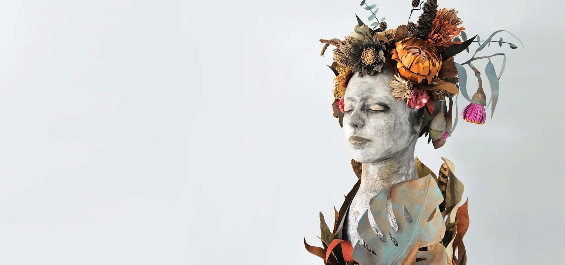 A sculpture of a woman with closed eyes, painted in gray tones, adorned with a crown of autumn paper leaves and flowers