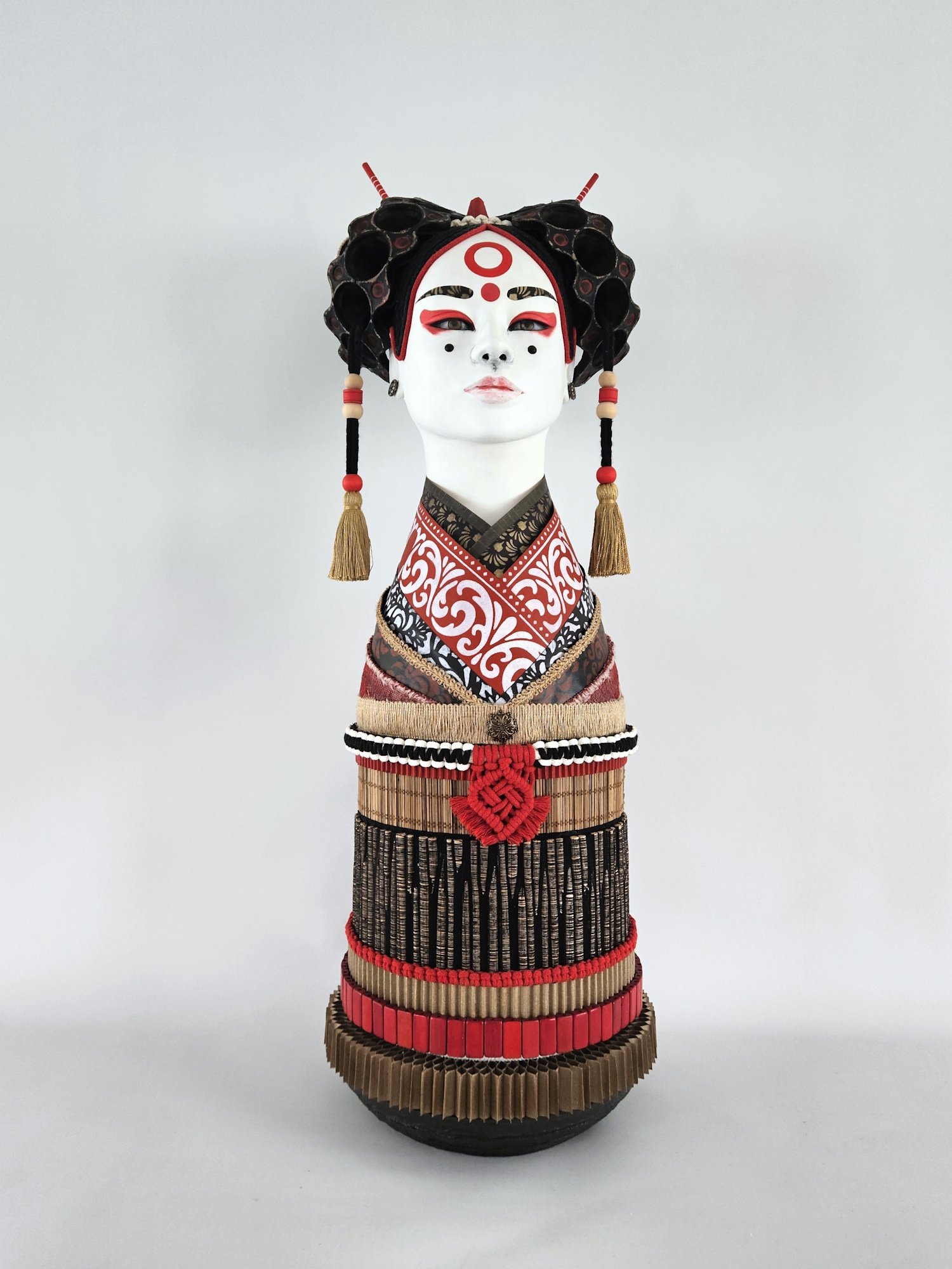 A traditional Japanes sculpture with a white face, red and black makeup, and an ornate headdress with black, red, and gold accents, wearing a red, black, and white kimono with intricate patterns, against a plain white background.