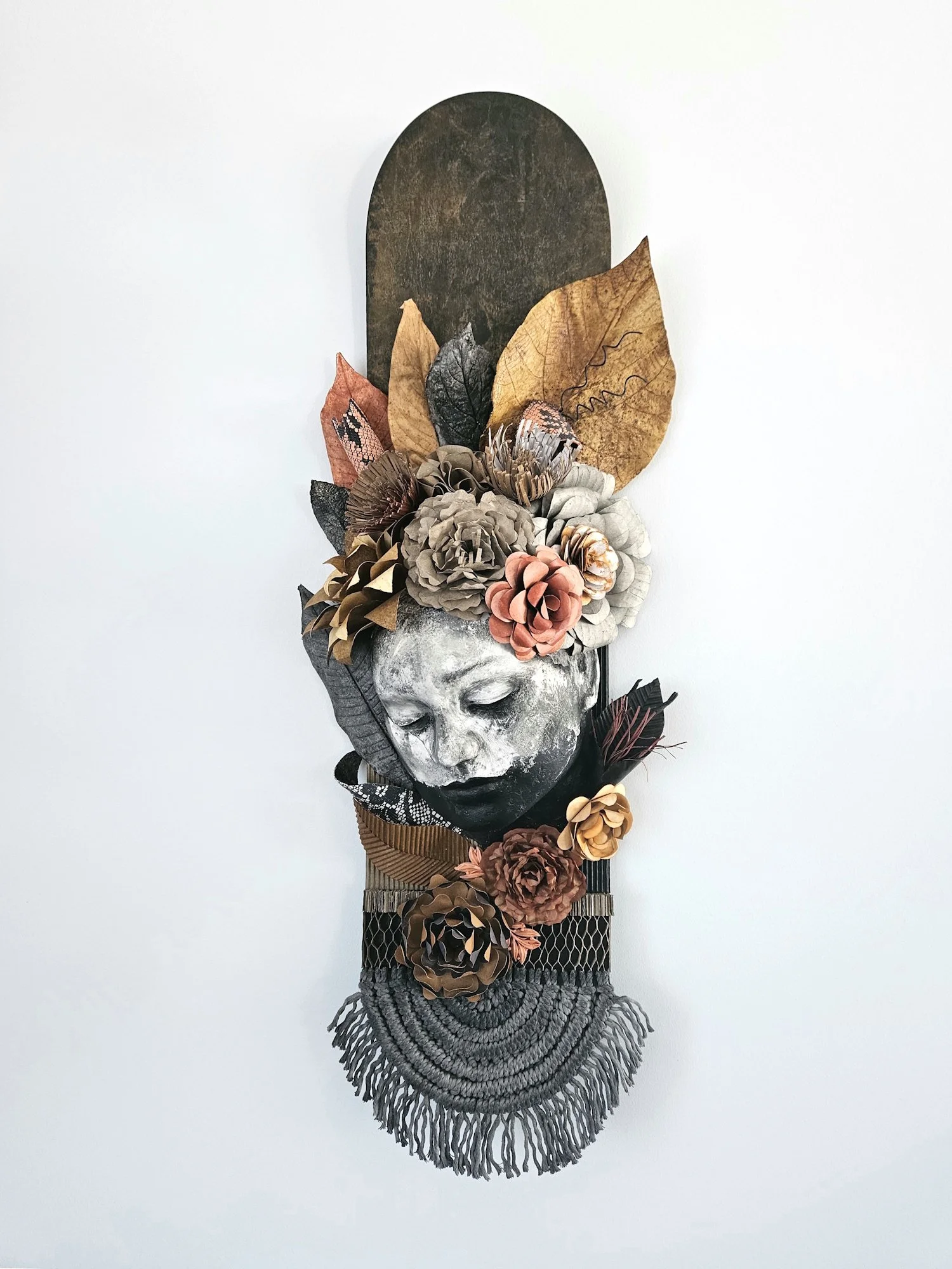 Mixed media art piece featuring a sculpted face with closed eyes, adorned with paper flowers and leaves, with a large arched wood background and layered materials including fabric, paper, and metal at the bottom.