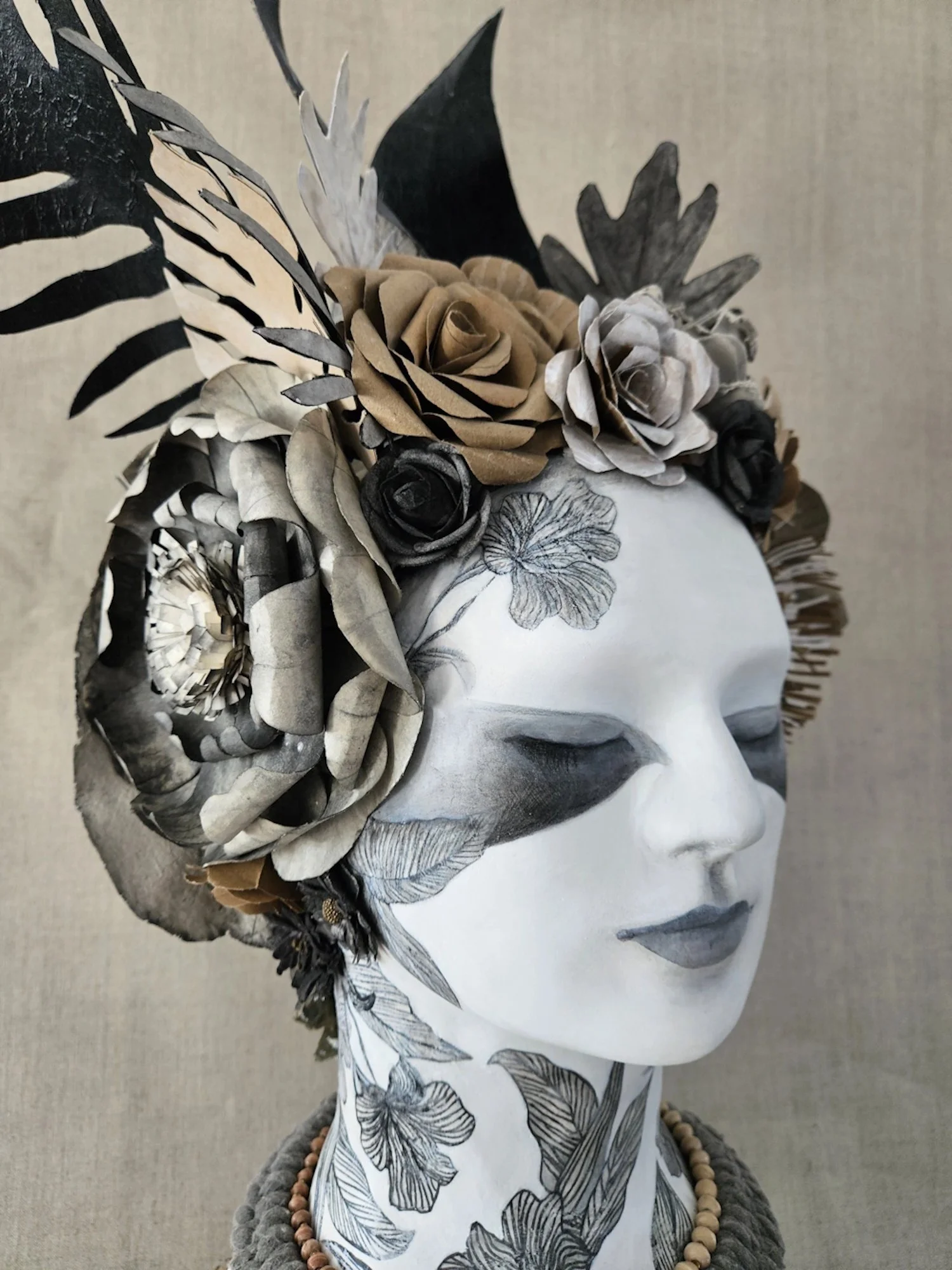A head sculpture with a floral headdress made of paper flowers and leaves, dressed in a botanical patterned fabric, with a beaded necklace, against a neutral background.