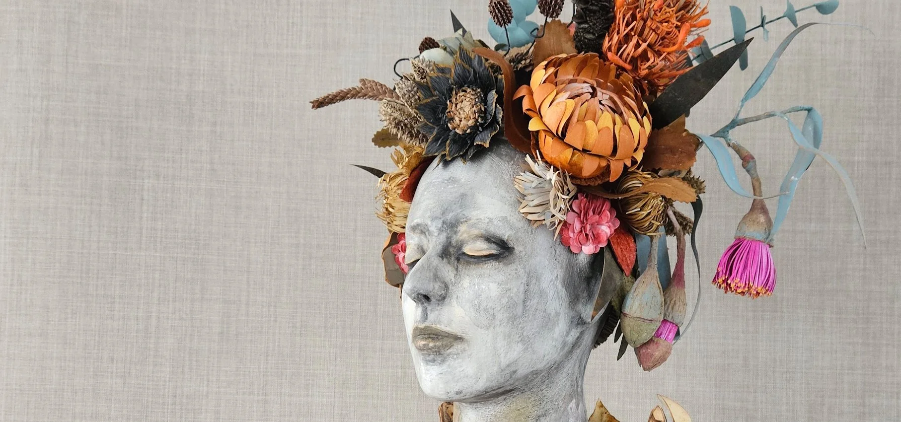 A sculpture of a human head with closed eyes, painted in a distressed white finish, topped with a large arrangement of paper flowers and foliage in shades of orange, pink, brown, and blue.