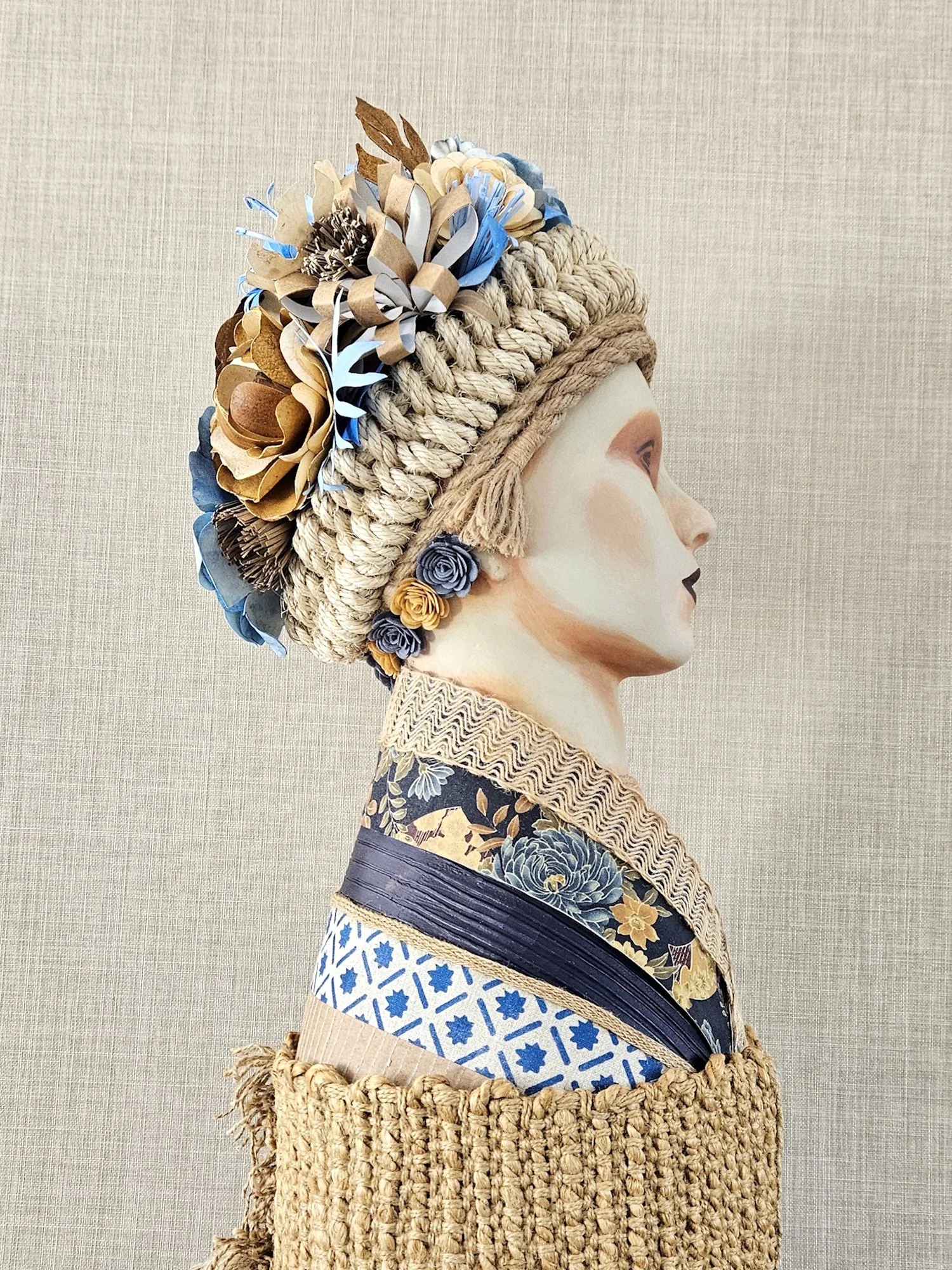 A sculpture of a bust wearing a Slavic dress with a wide woven straw hat decorated with embroidered flowers, paper flowers, and ribbon details against a plain light-colored background.