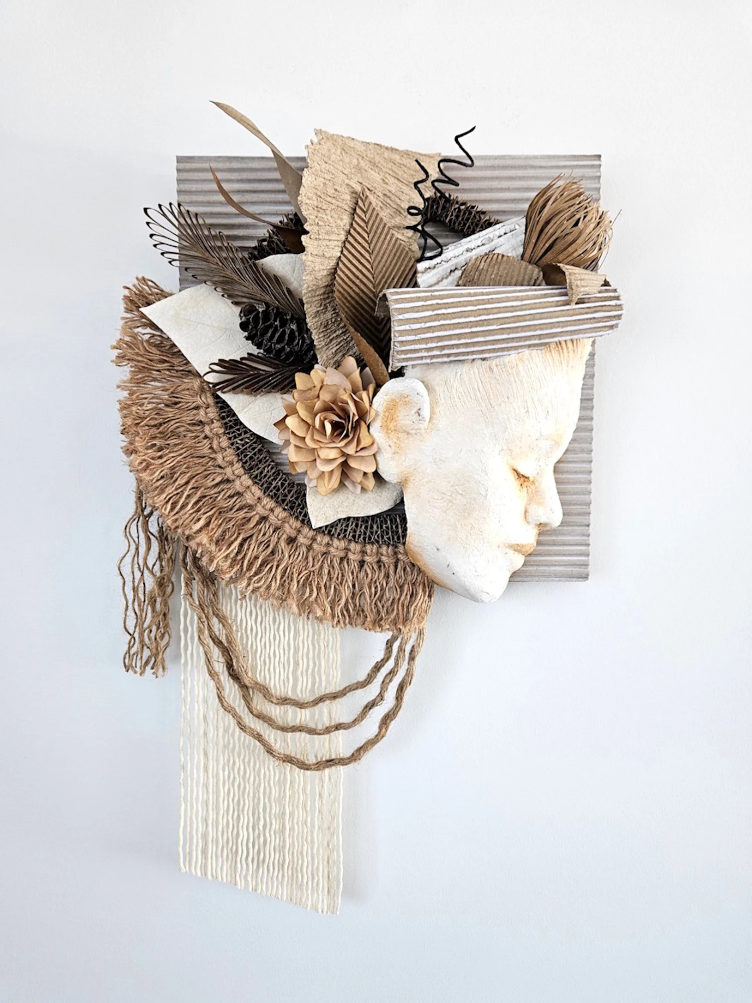 Mixed media art piece with a face profile, combining natural elements like paper flowers and leaves, and textured materials arranged on a wall.