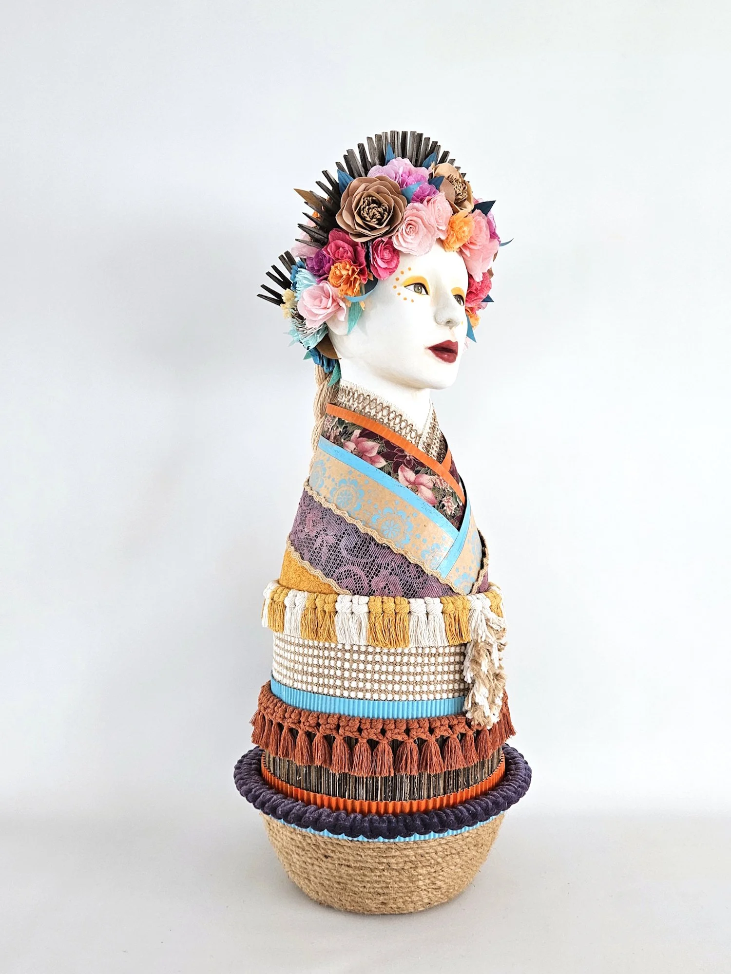 A sculpture of a woman with a face painted white, adorned with a colorful headdress of paper flowers, and dressed in layered, patterned fabric with decorative trims, set against a plain light background.