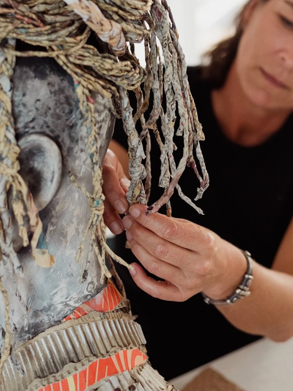 An artist working on a textured mixed-media art piece  covered in various layers of cardboard and paper, with hands adjusting or attaching a detail.