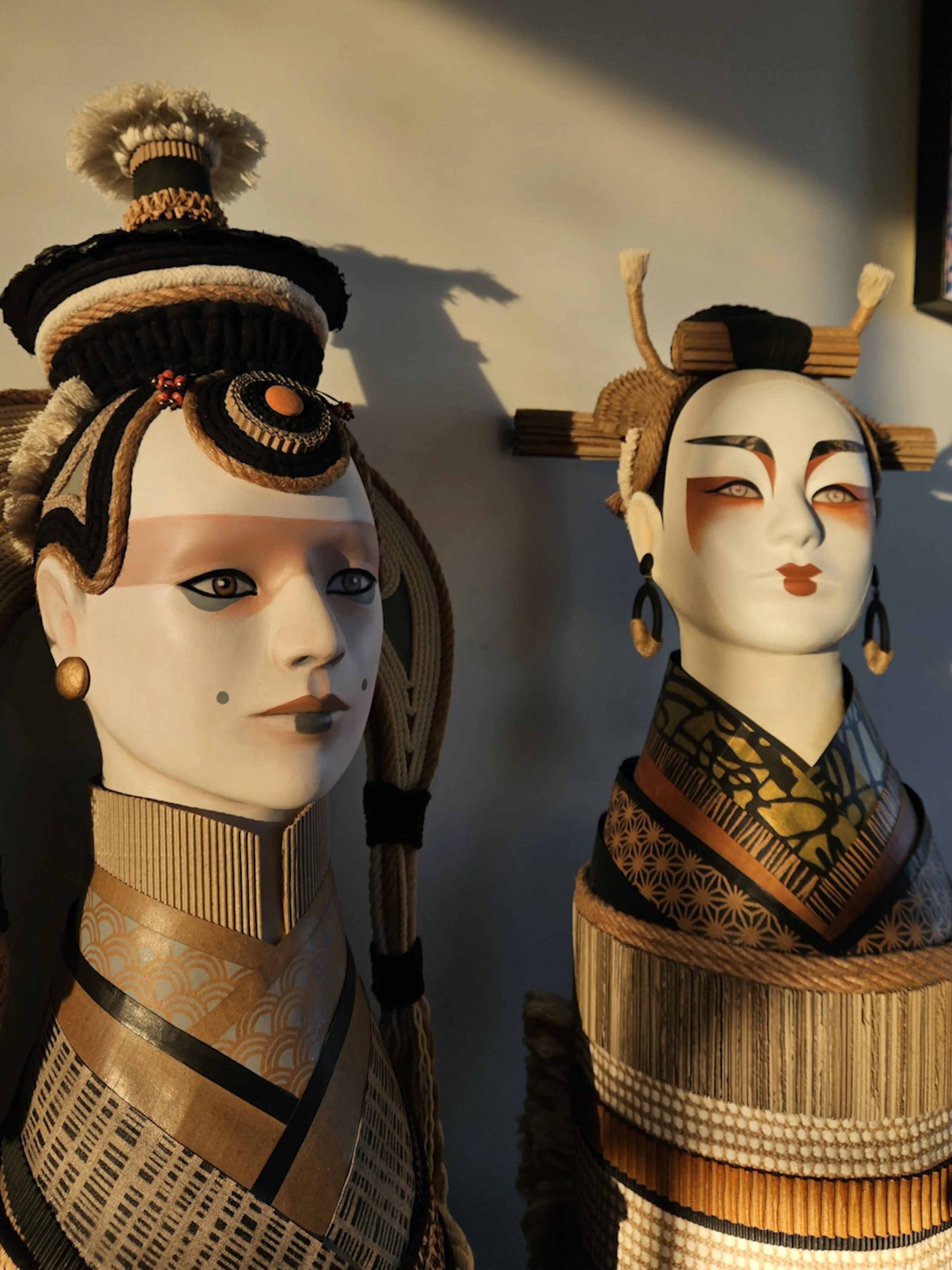 Two traditional Asian masks with elaborate headpieces and costumes, displayed on stands against a plain wall.